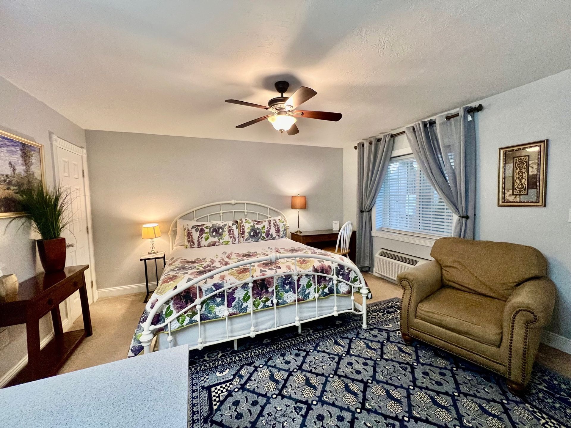 Bedroom with a white metal bed, blue patterned rug, armchair, and curtains.