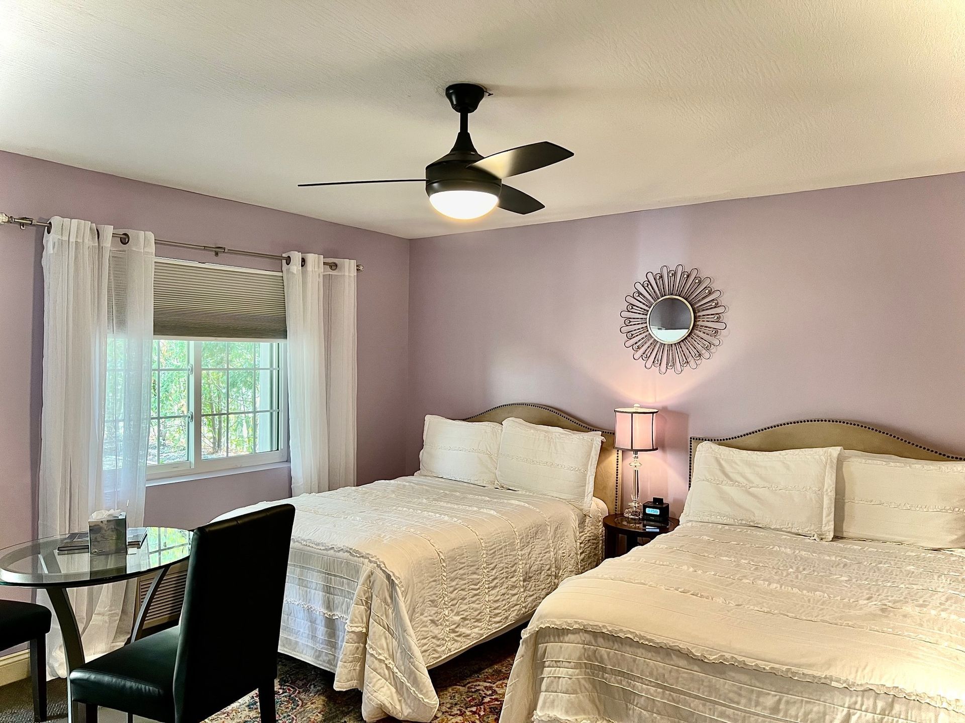 Bedroom with two beds, lavender walls, window, table, ceiling fan, and decorative mirror.