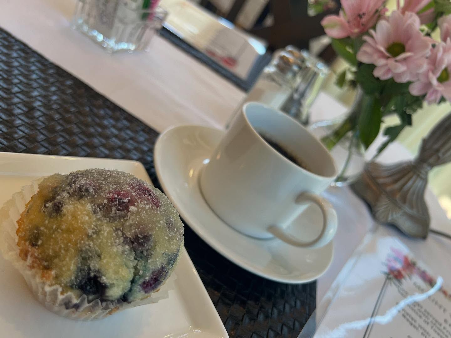 Muffin with sugared top, coffee in white cup and saucer, and pink flowers on a table.