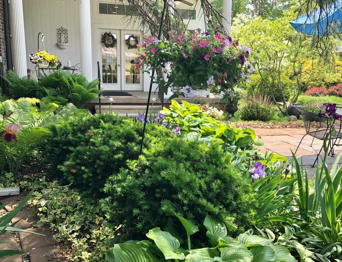 Exterior of white building with flower-filled garden. Hanging basket of pink flowers. Green foliage.