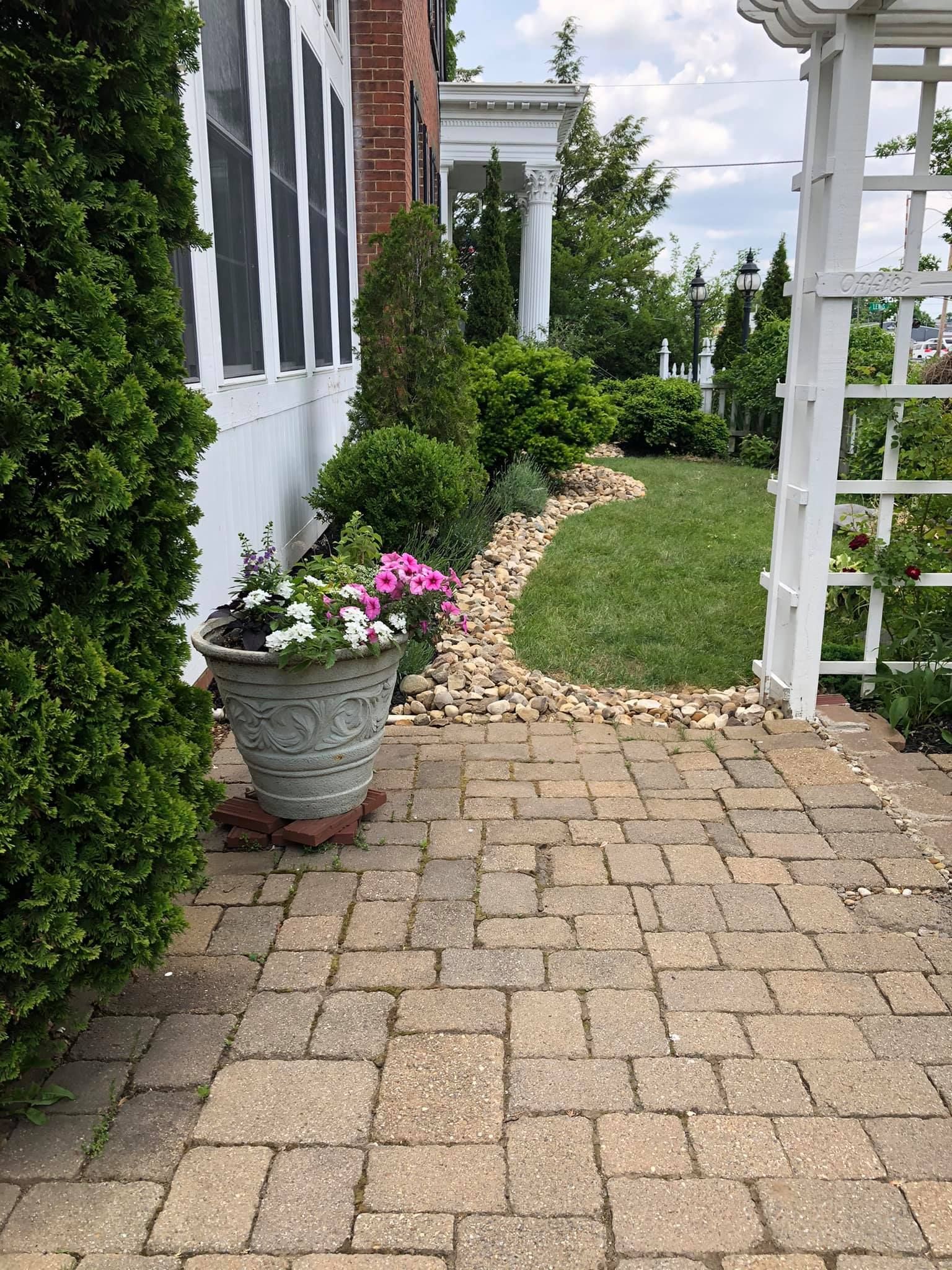 Brick walkway leading to a garden with a stone-lined flower bed, green grass, and a white trellis.