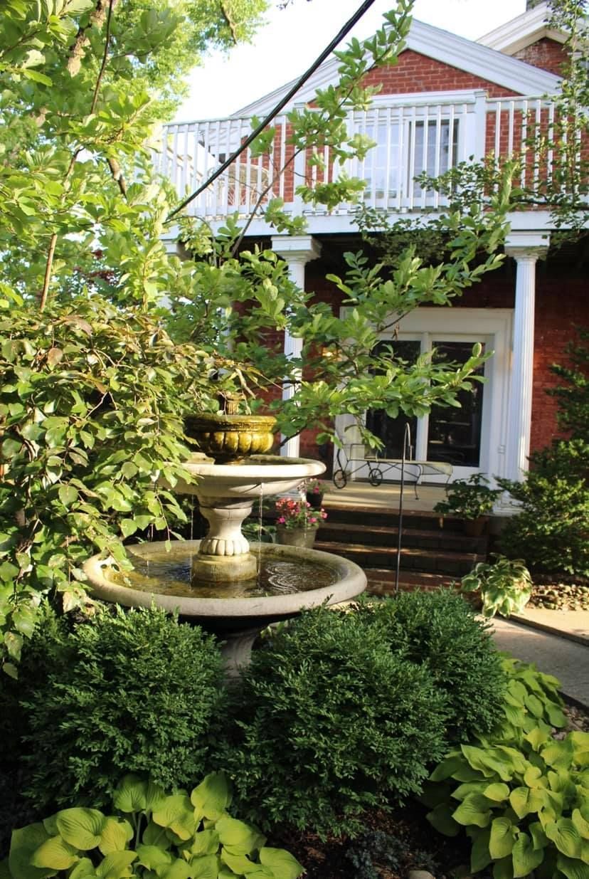 Stone fountain in a lush garden, with red brick house and white balcony in the background.