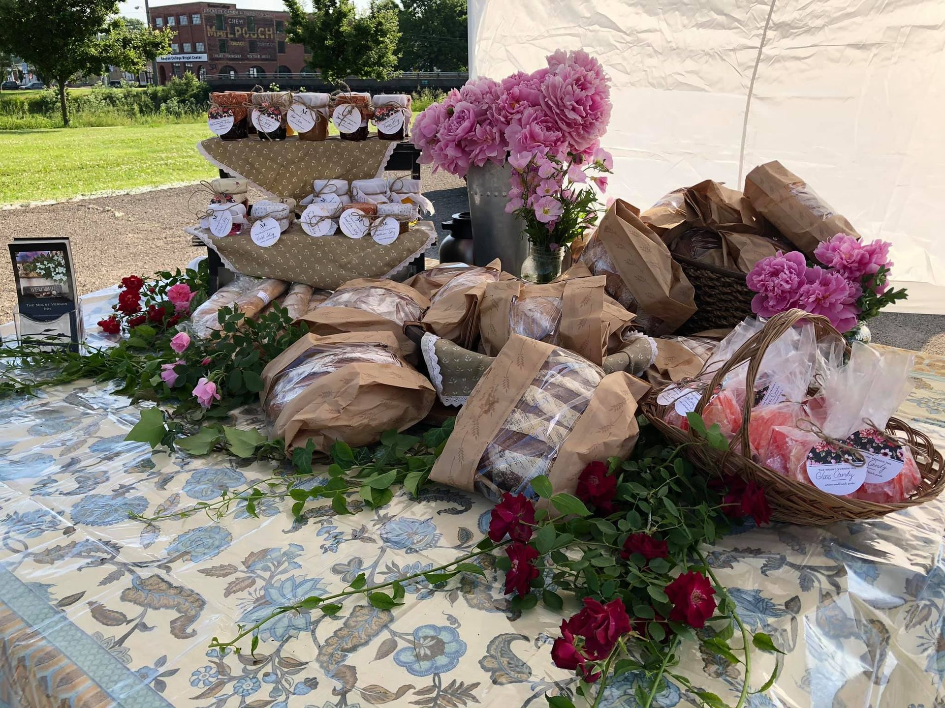 Farmers market display: baked goods, jams, flowers on a table with a patterned tablecloth.
