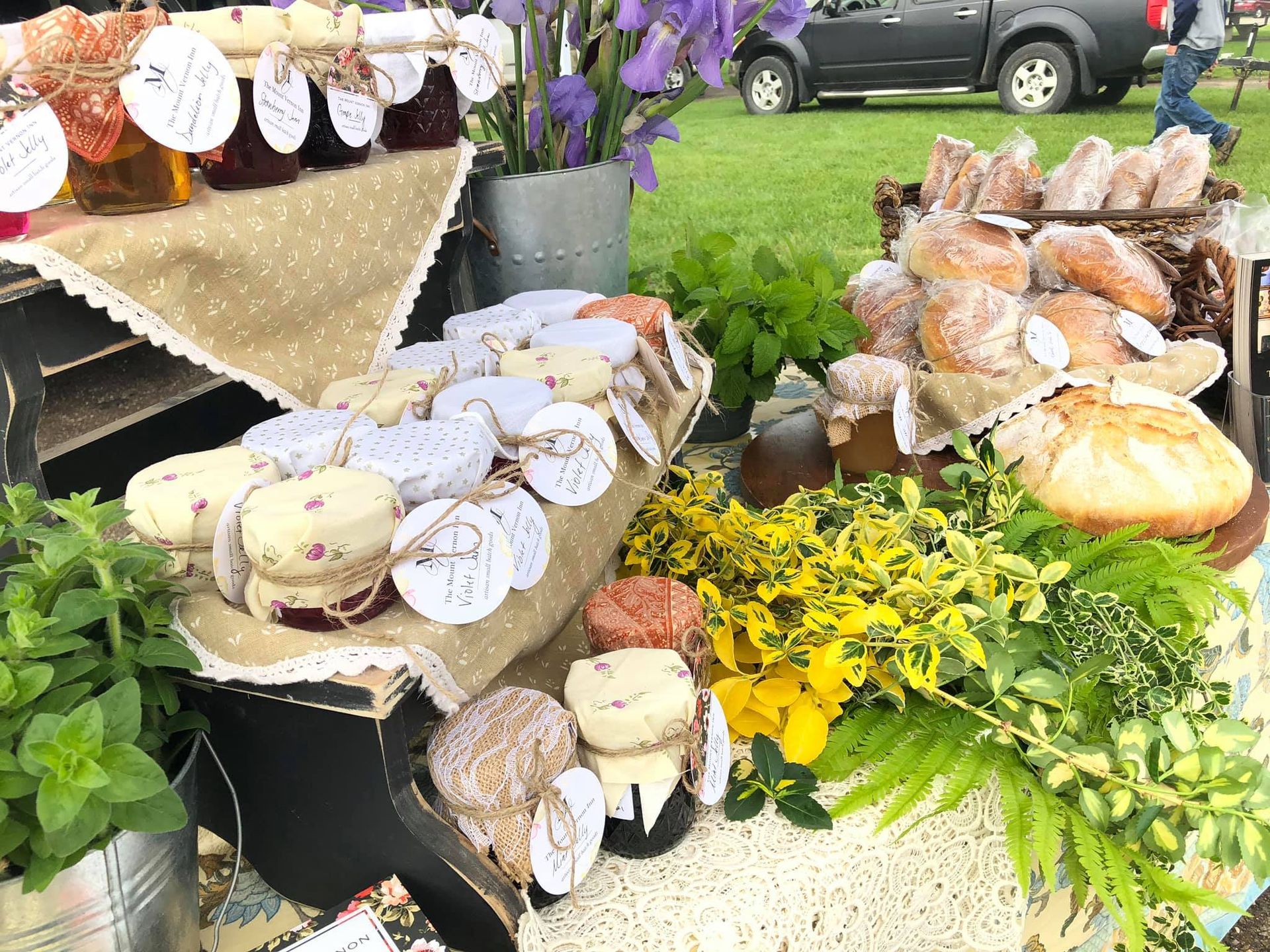 Market stall display of preserves, baked goods, and flowers, outdoors. Jars of jelly, round pastries, and bread.