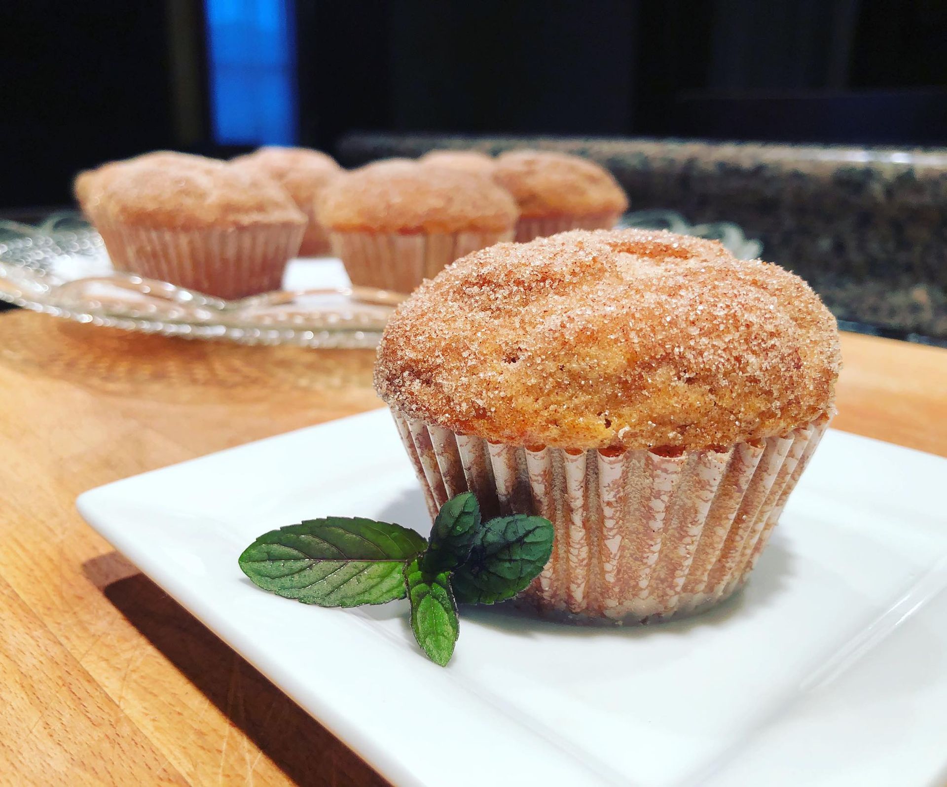 A muffin sprinkled with cinnamon and sugar, garnished with mint, on a white plate. Other muffins in the background.