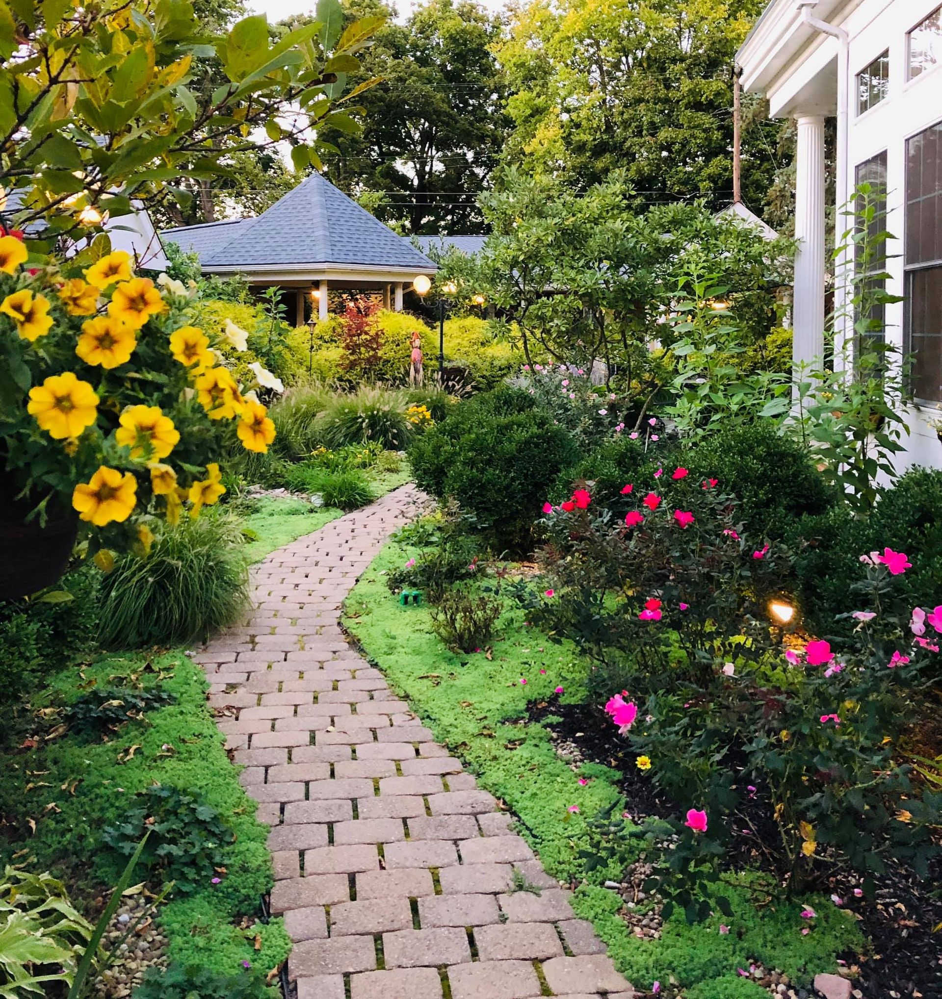 Brick pathway through a lush garden with yellow and pink flowers, leading to a house.