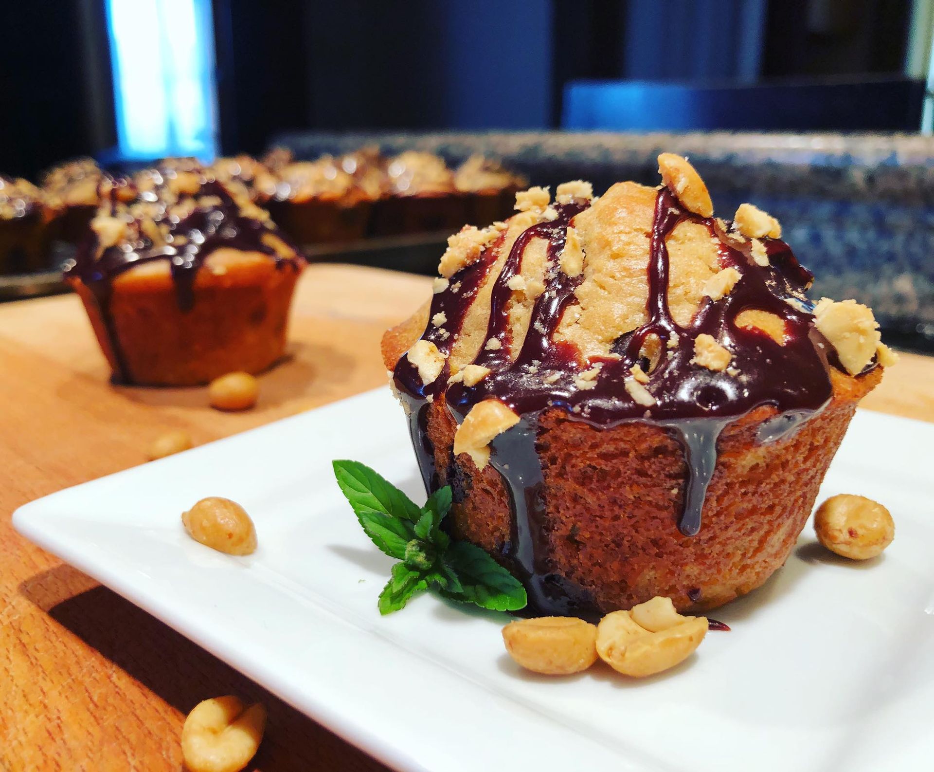 Muffin topped with chocolate drizzle, crushed nuts, and mint on a white plate with peanuts scattered.