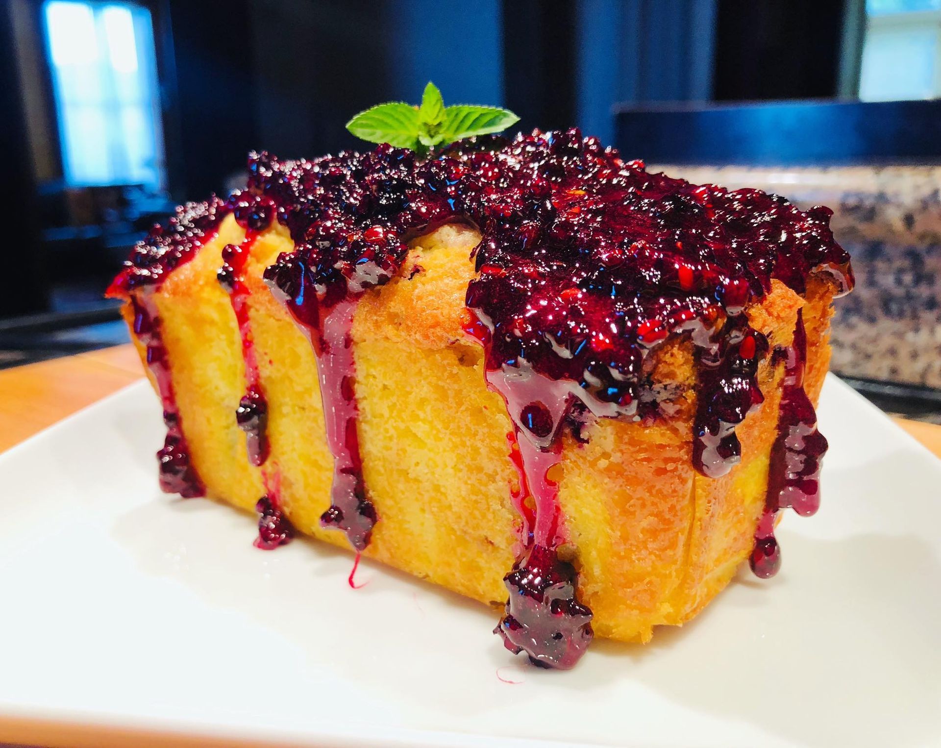 Golden pound cake with dripping blackberry compote, topped with a mint sprig, on a white plate.