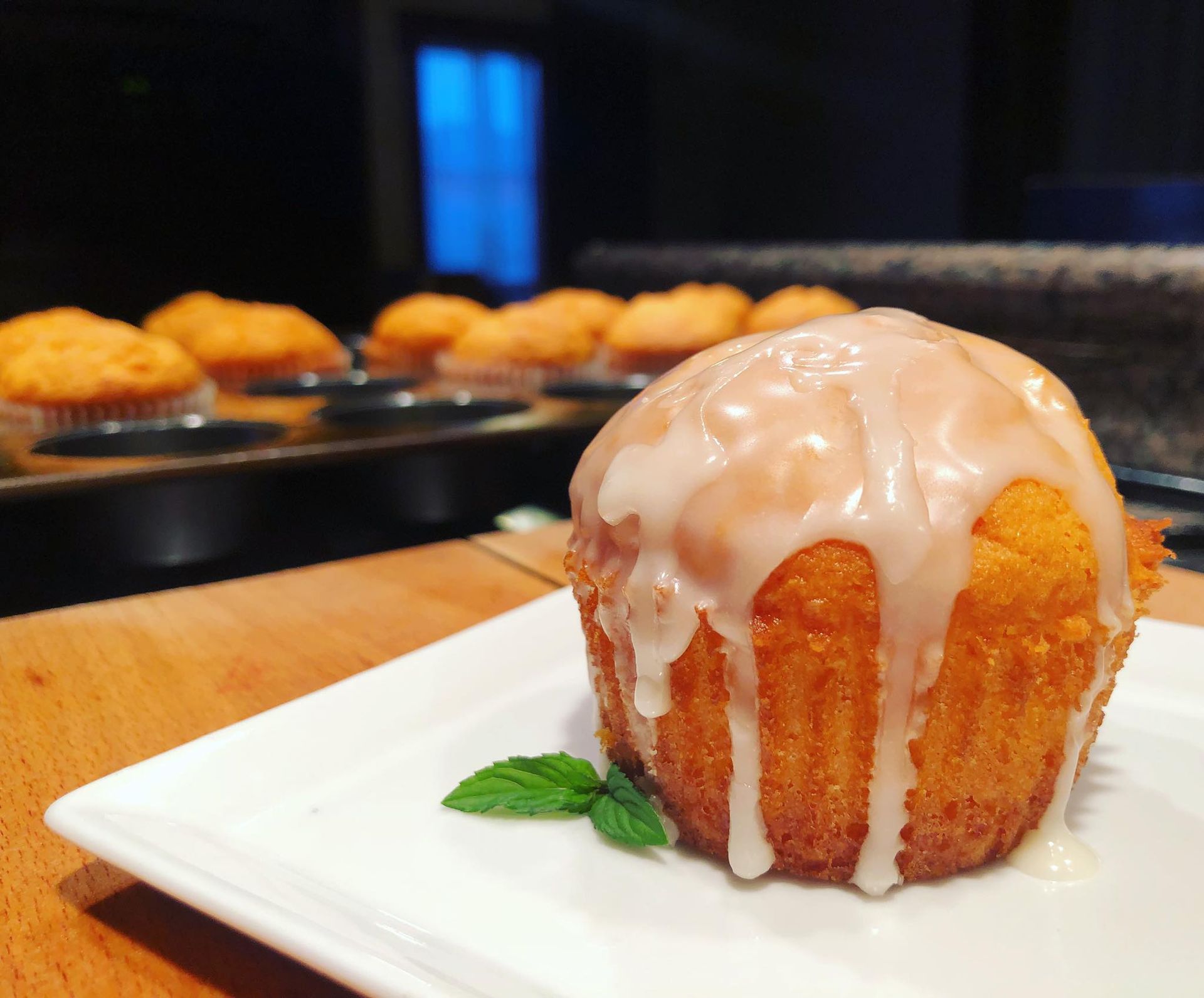 Muffin with glaze on a white plate, mint garnish; muffins in a baking tray in the blurred background.