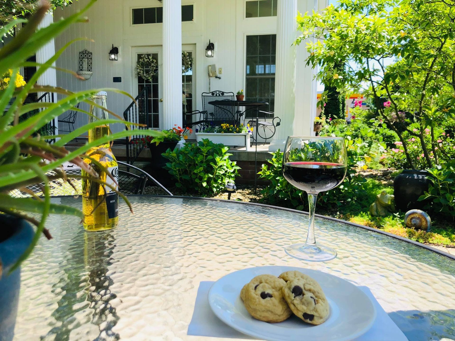 Glass table with wine and cookies. White house with porch in the background, bottle of beer and plants.