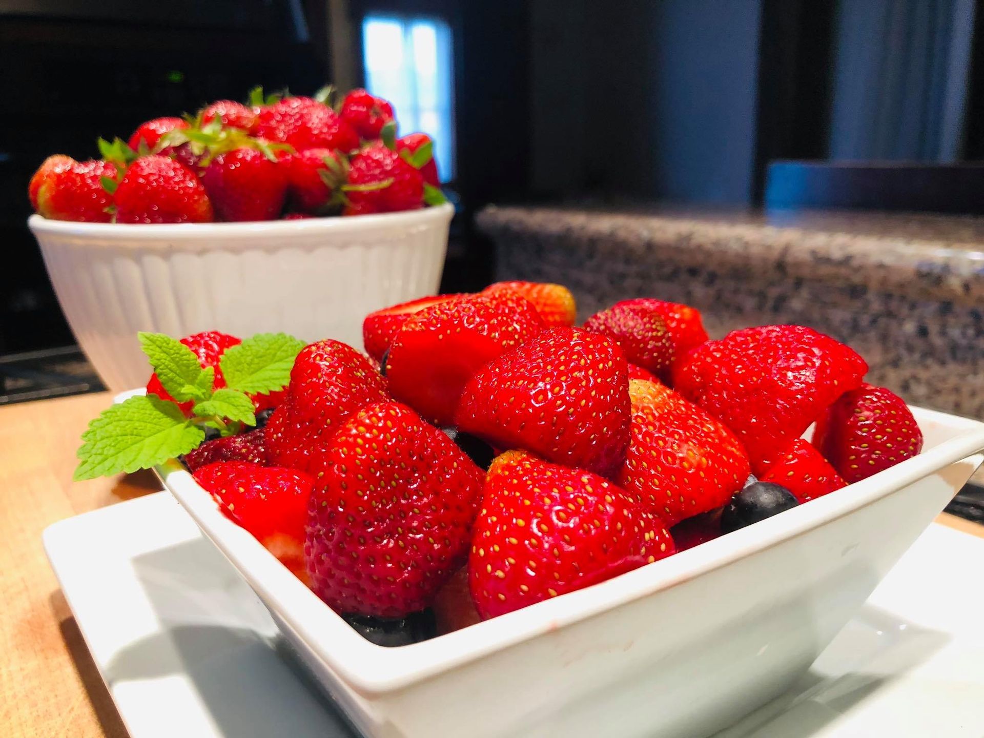 Two white bowls overflowing with ripe red strawberries, with a sprig of mint.