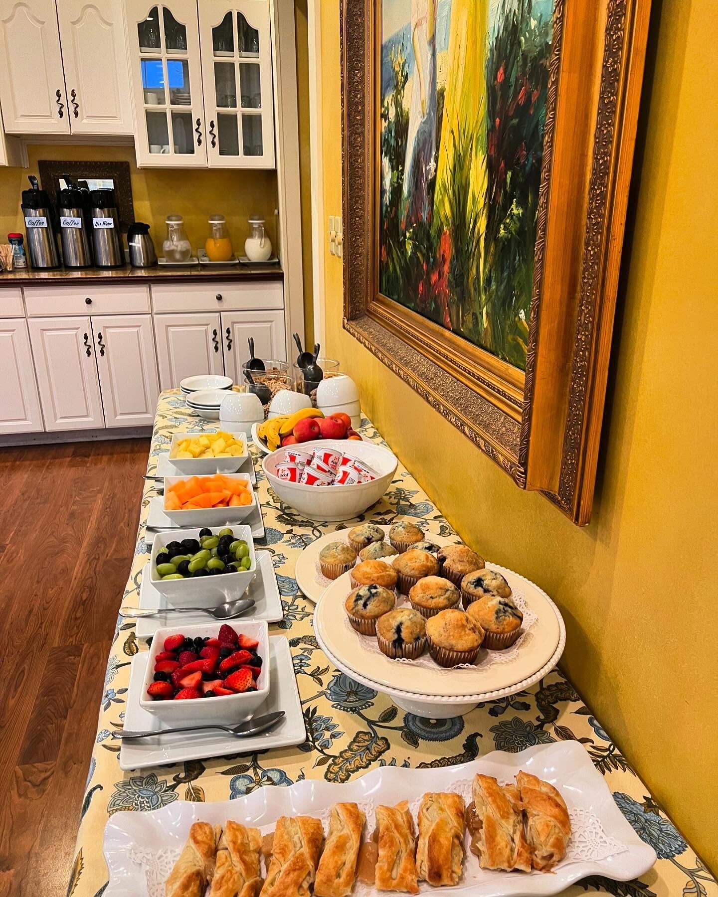 Breakfast buffet on a yellow table with muffins, pastries, fruit, and yogurt near a painting and white cabinets.