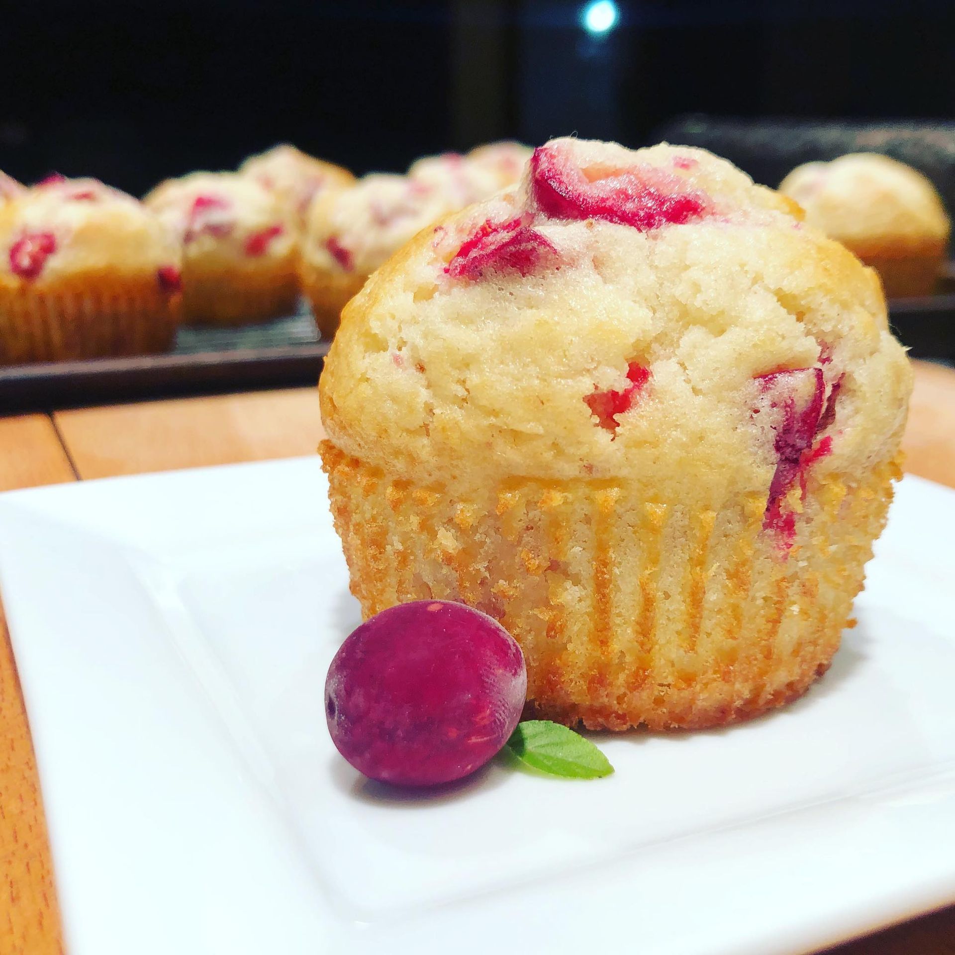 Cranberry muffin on white plate with a cranberry, other muffins blurred in the background.
