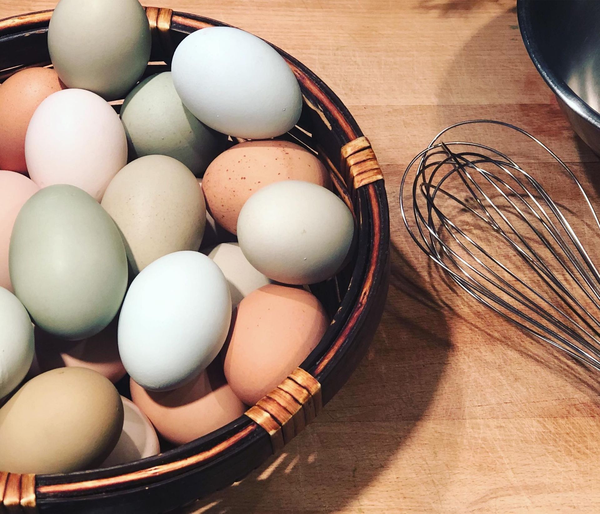 Basket of colorful eggs and a whisk on a wooden surface.
