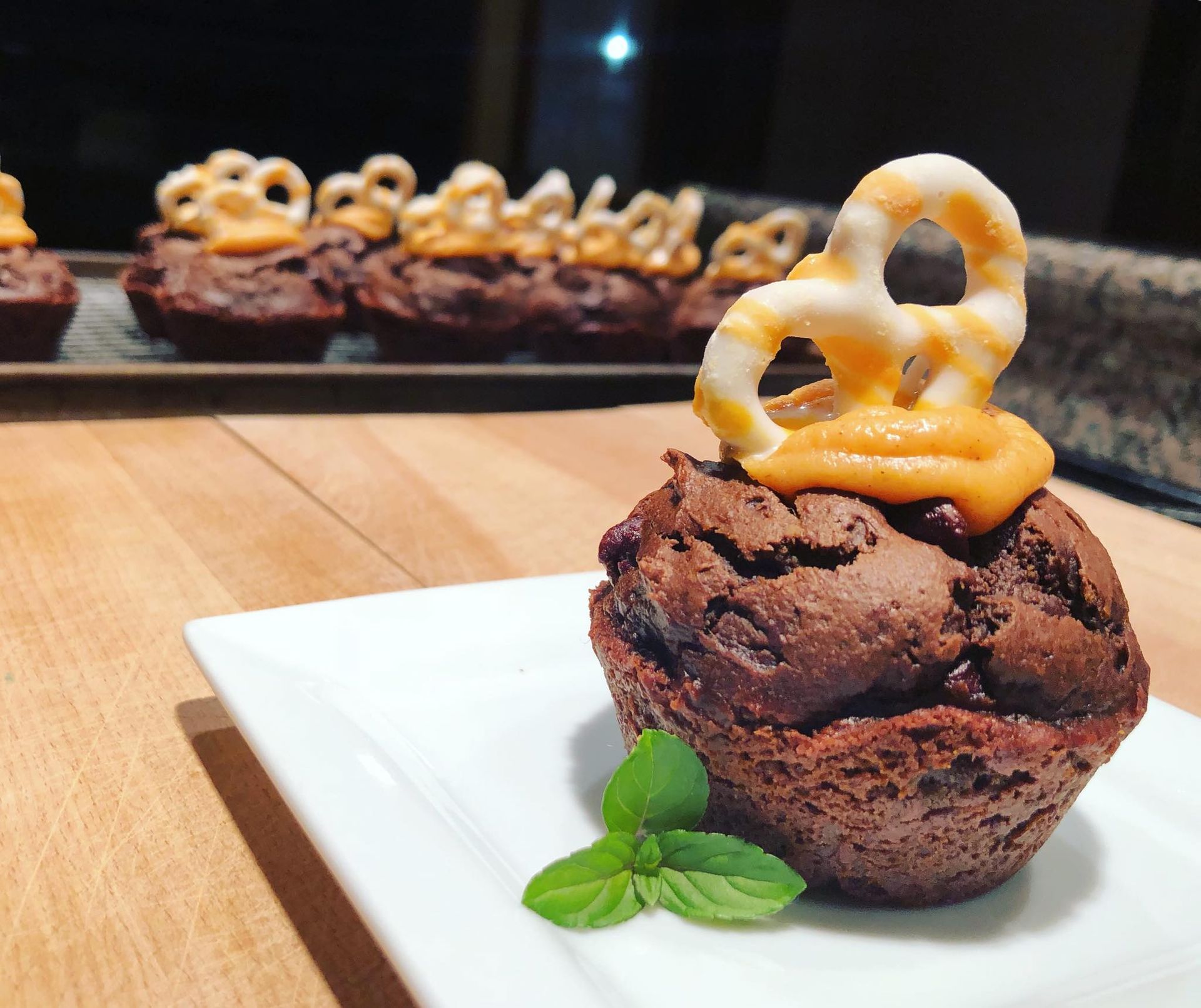 Chocolate muffin topped with caramel and a pretzel, mint sprig on the plate.