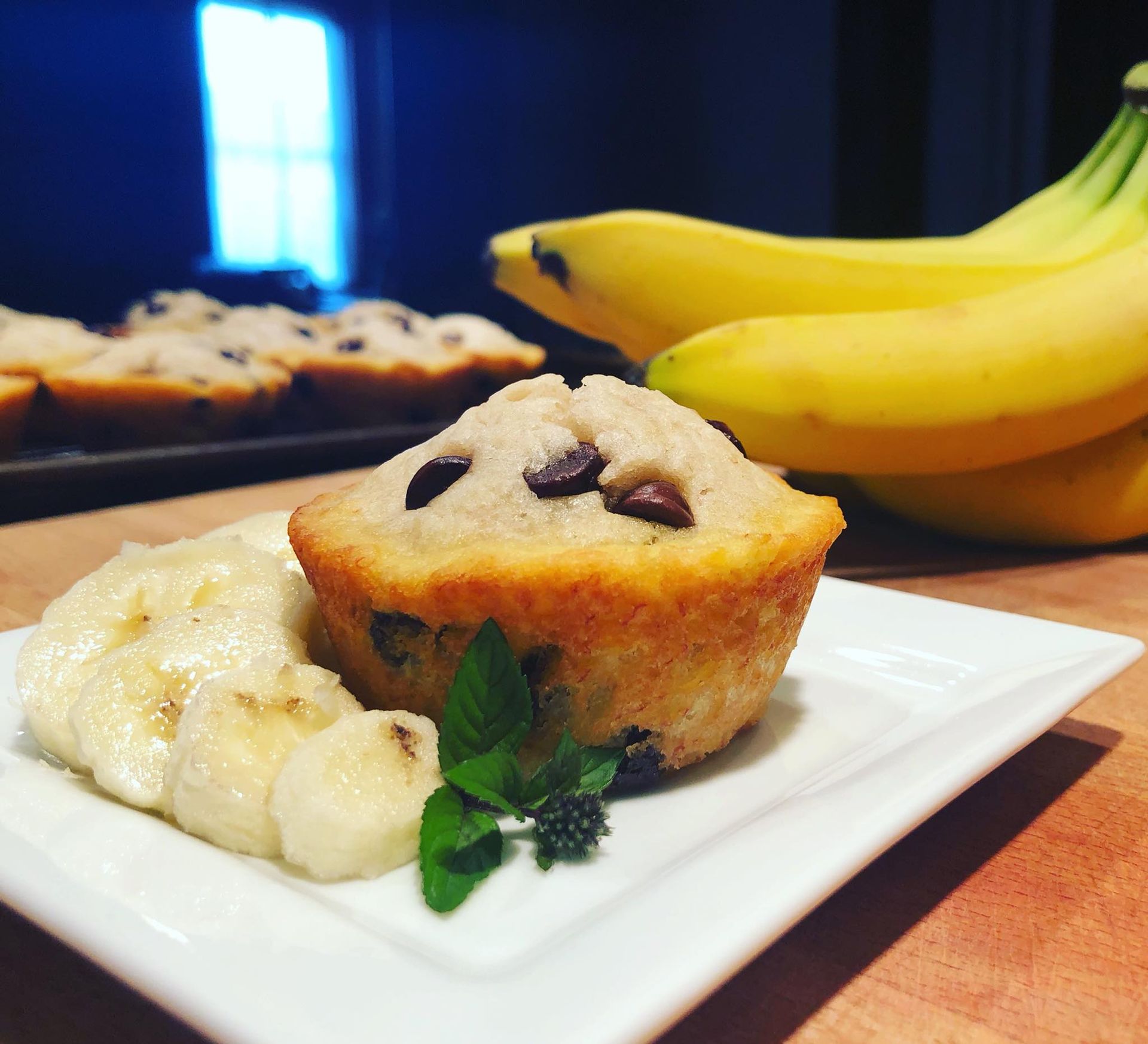 Banana chocolate chip muffin on a plate with banana slices and a sprig of mint; bananas in the background.