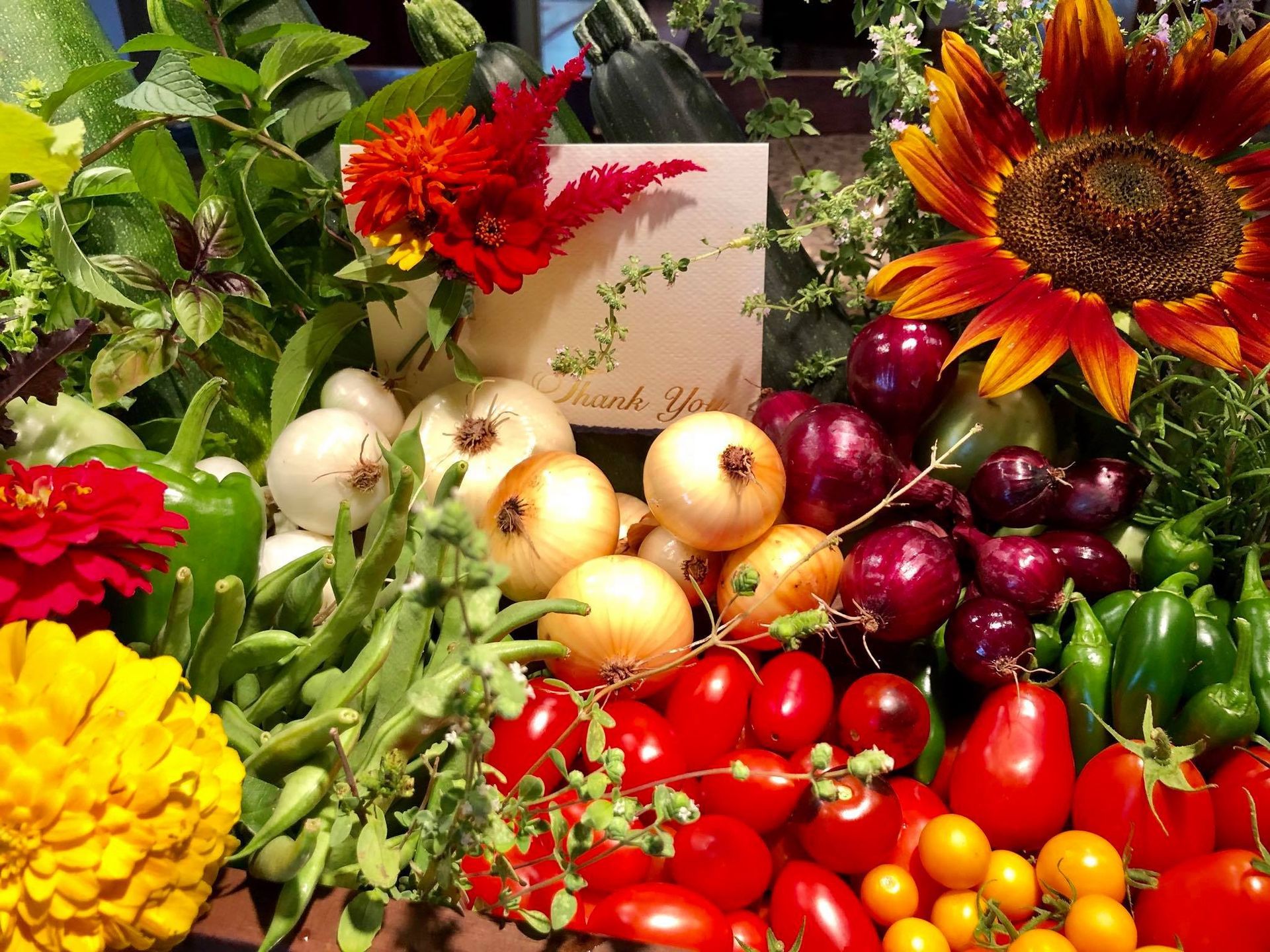 Assortment of colorful vegetables and flowers: onions, tomatoes, peppers, sunflower, and red celosia.