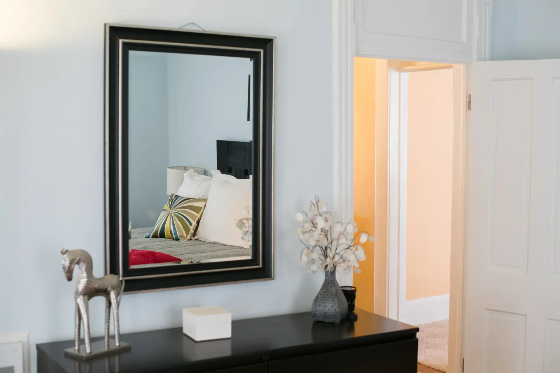Black dresser with mirror reflecting bedroom. Silver horse statue and vase with white flowers. Light blue walls.