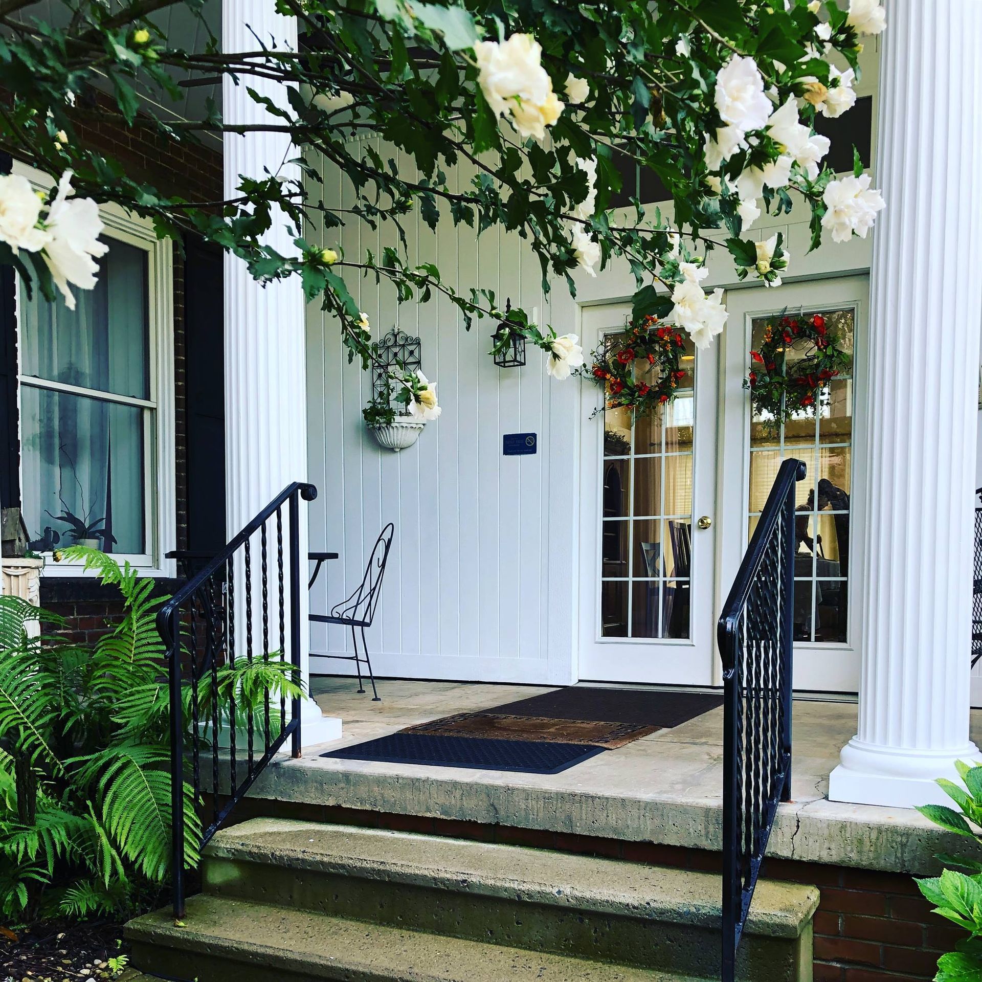 White-columned porch with steps, black railing, double glass doors with wreaths, flowering tree branches, and a fern.