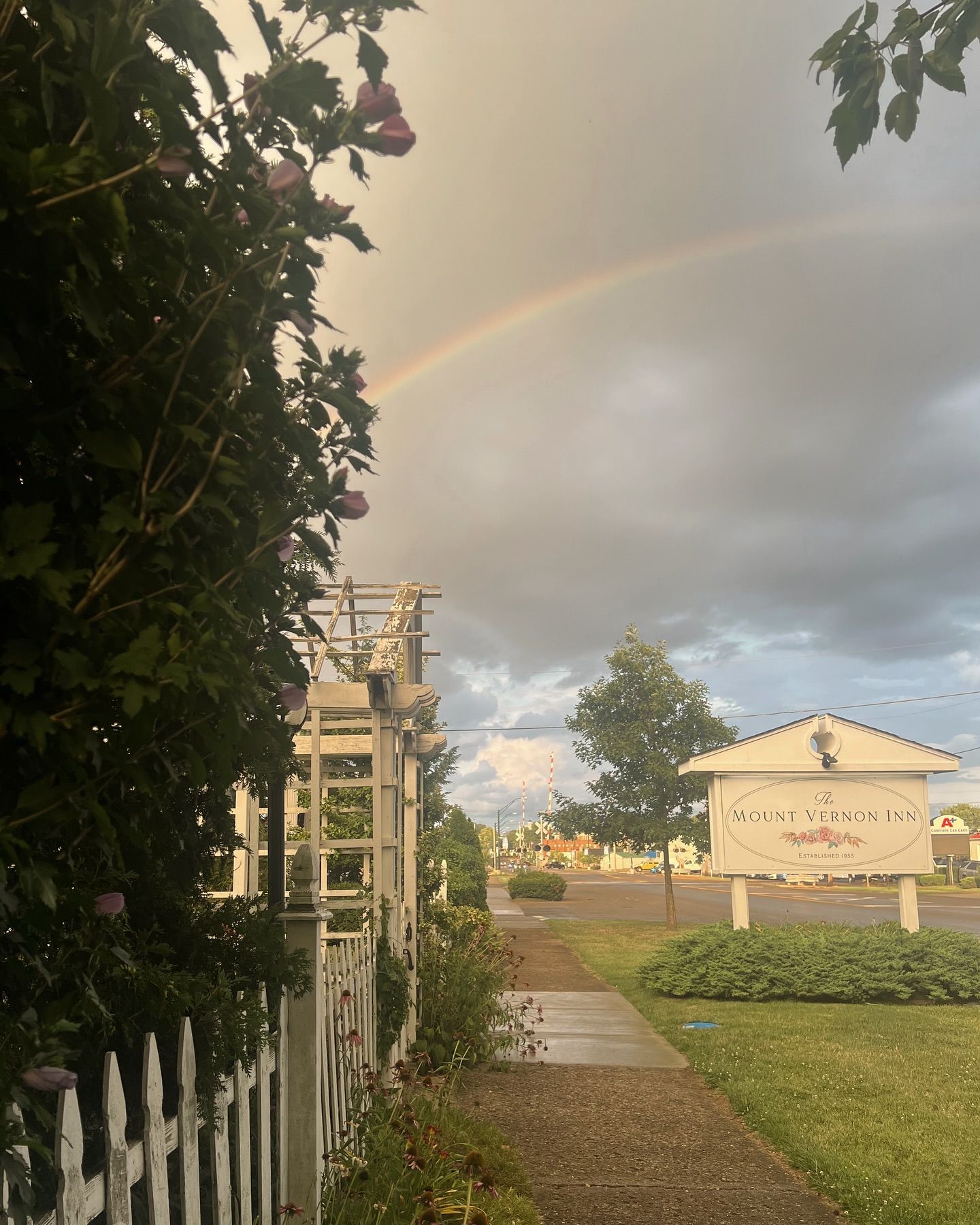 Rainbow arches over a street with a sign, bushes, a sidewalk, and a white fence.
