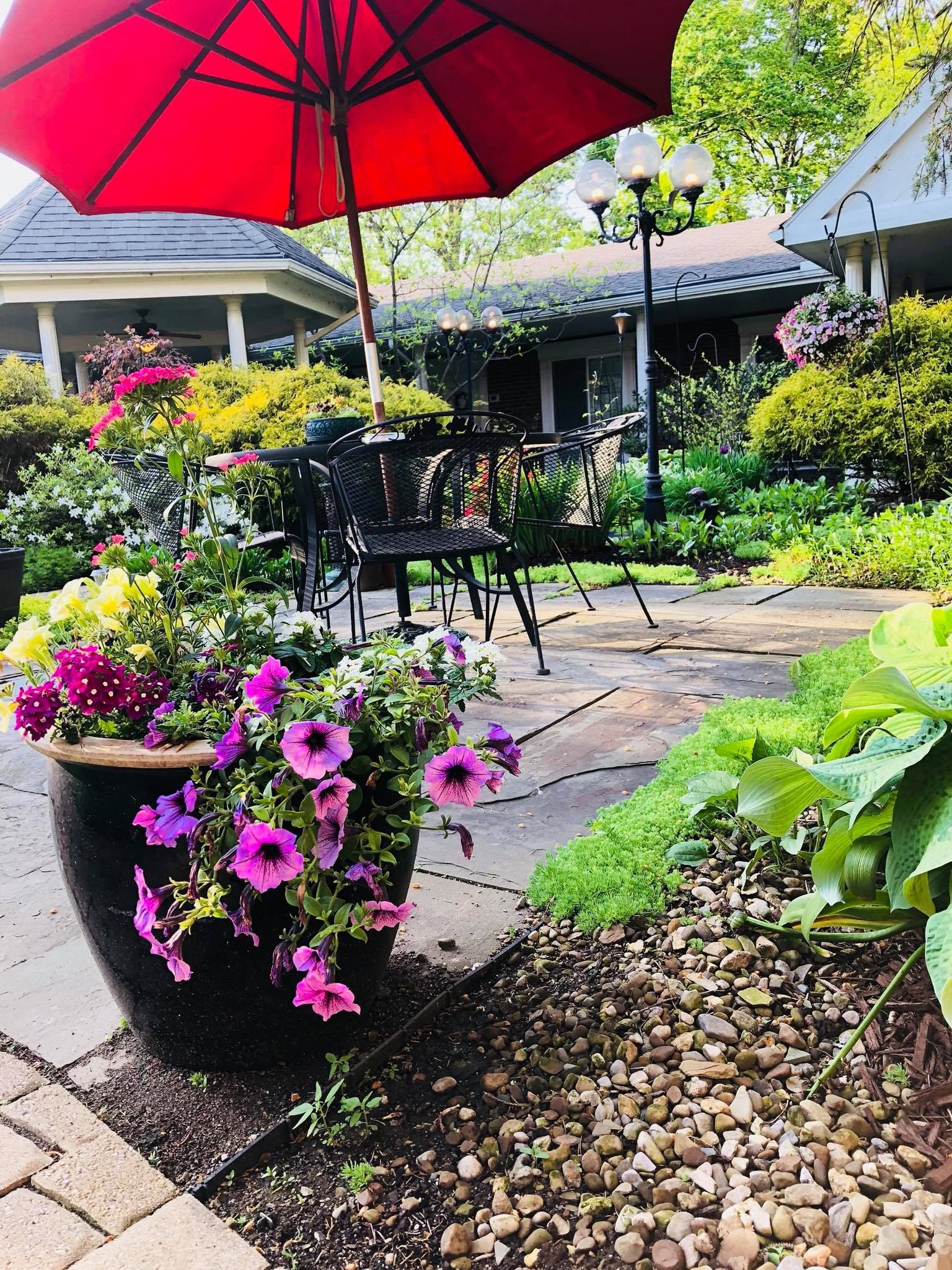 Red umbrella shades patio with potted flowers, dark metal table, and lush greenery.