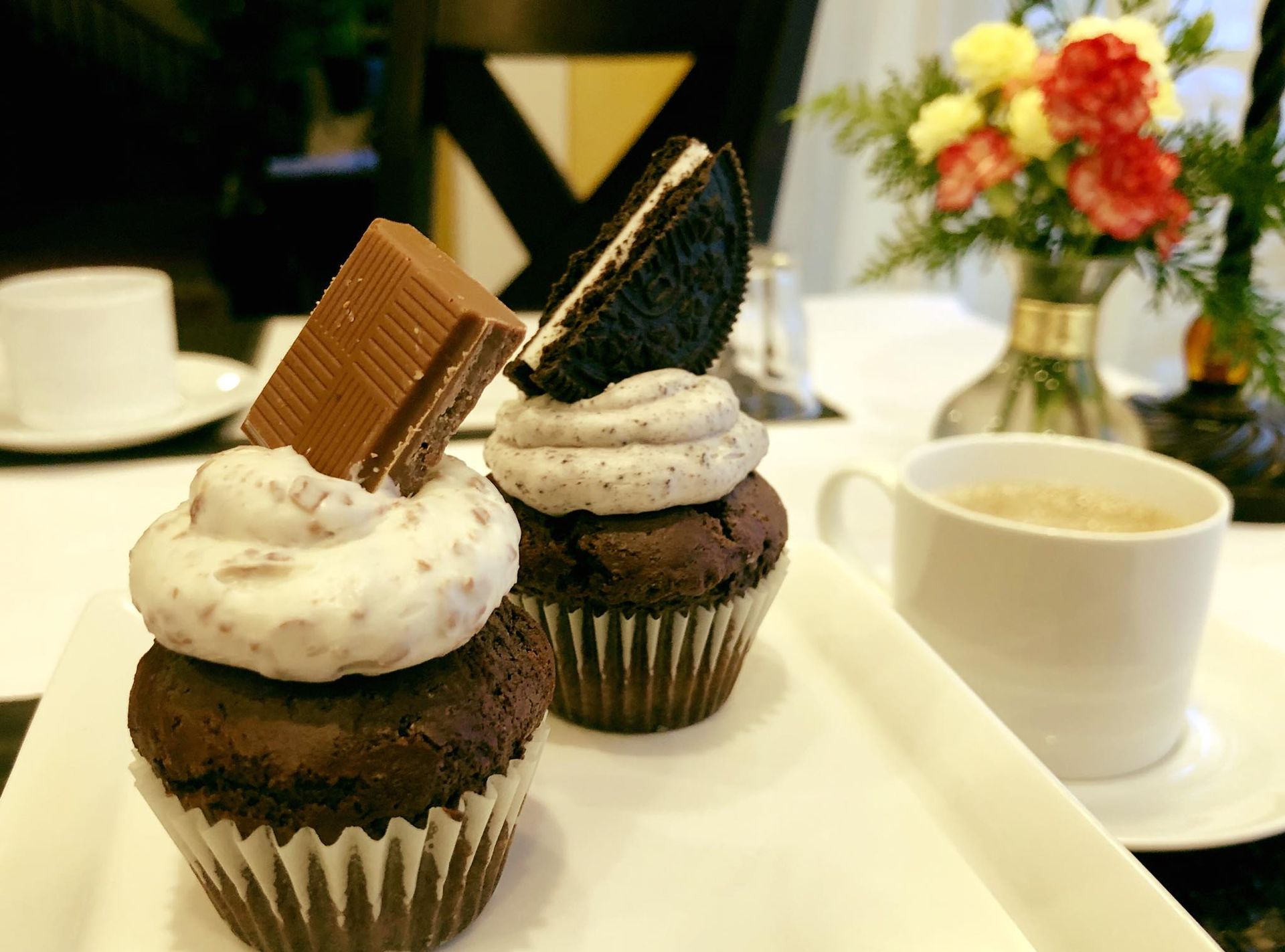 Two chocolate cupcakes with frosting, topped with a Kit Kat and an Oreo, beside a cup of coffee and flowers.