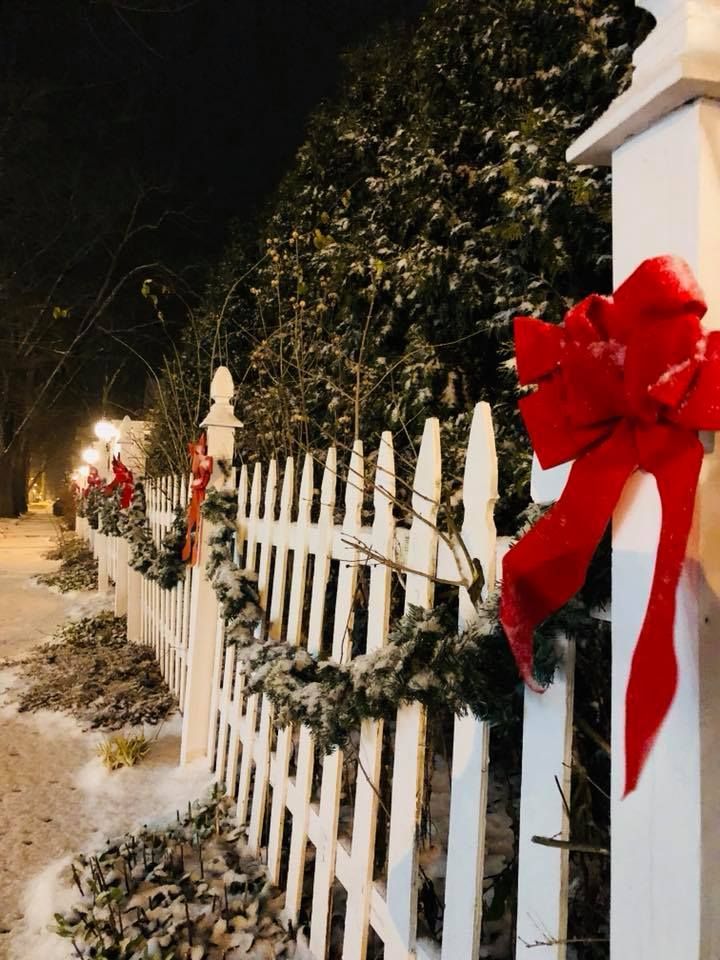 White picket fence decorated with red bows and garland in a snowy winter setting at night.