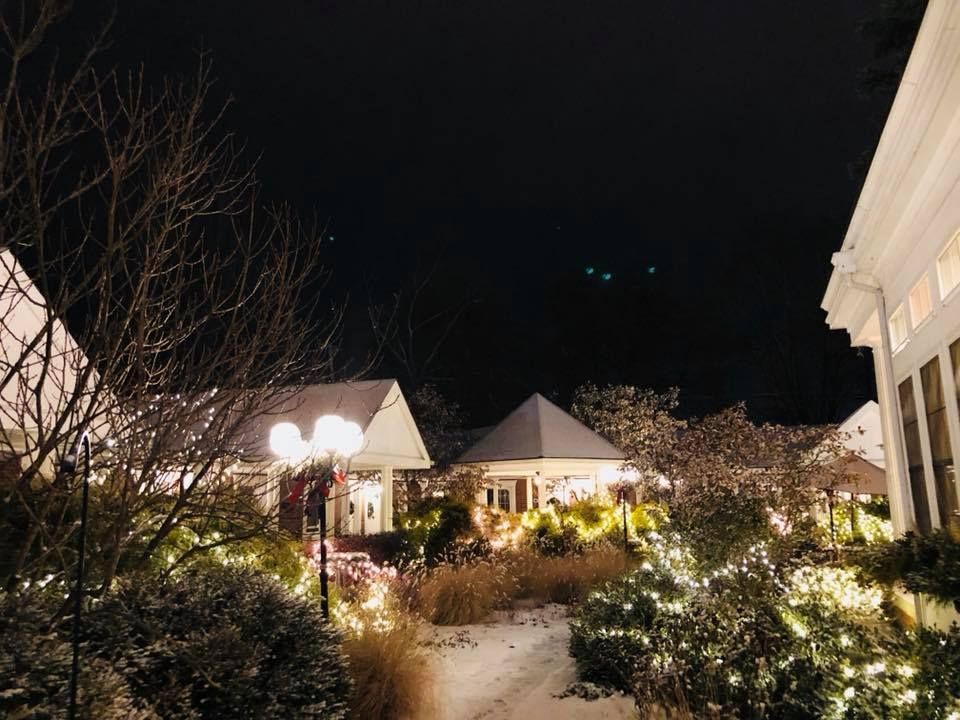Snow-covered outdoor scene at night, lit with string lights. Buildings and trees are visible against the dark sky.