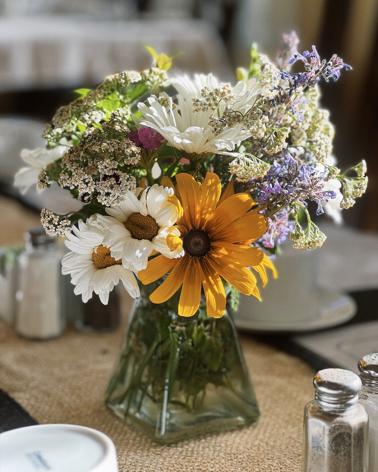 Bouquet of yellow, white, and purple flowers in a glass vase on a table with salt shakers.