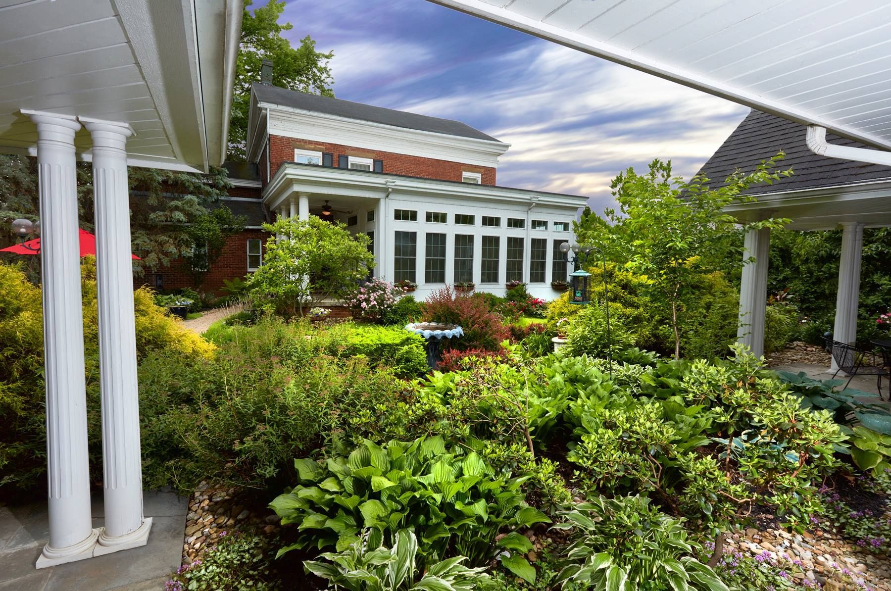 Lush garden with a brick house featuring a white porch and pillars; solar panels on a pergola.