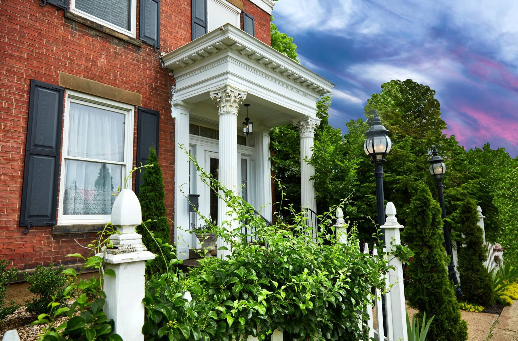 Brick house with white porch and trim, black shutters, white picket fence, green foliage, cloudy sky.