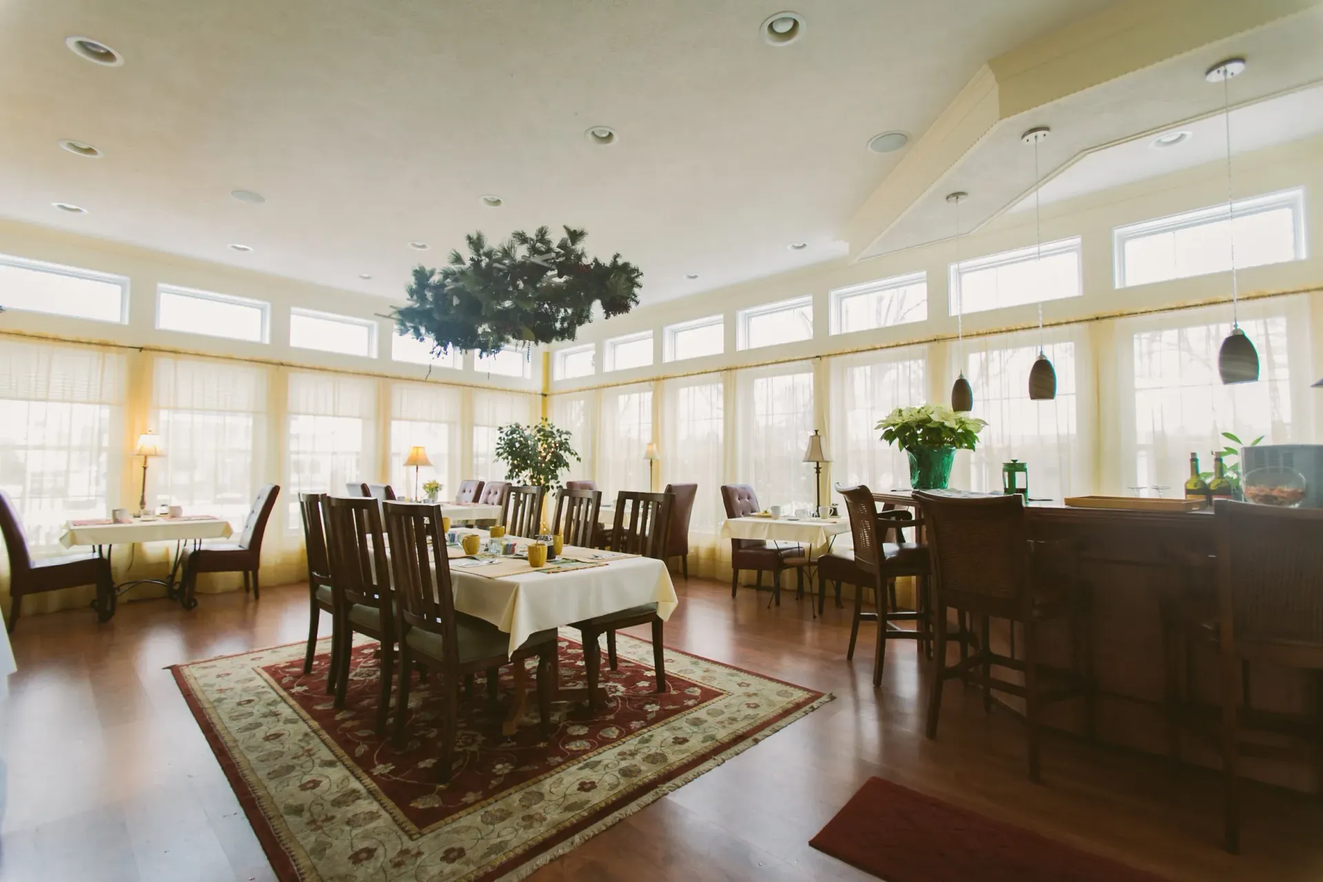 Interior of a dining room with tables set, a bar, and large windows with cream curtains.