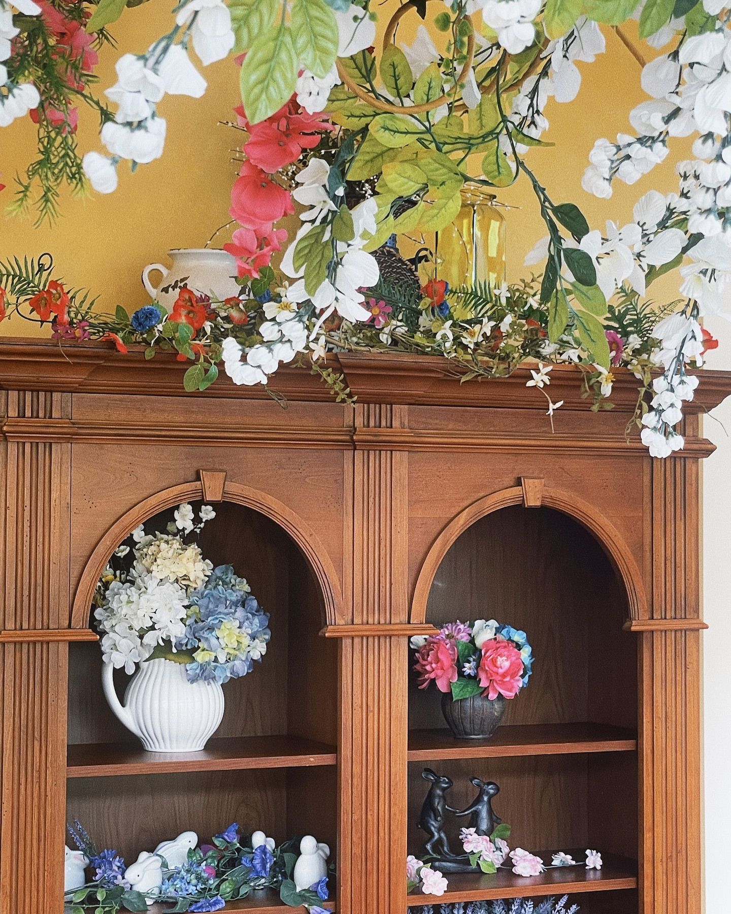 Wooden bookcase decorated with flowers, white and blue vase, colorful blooms on a yellow wall.