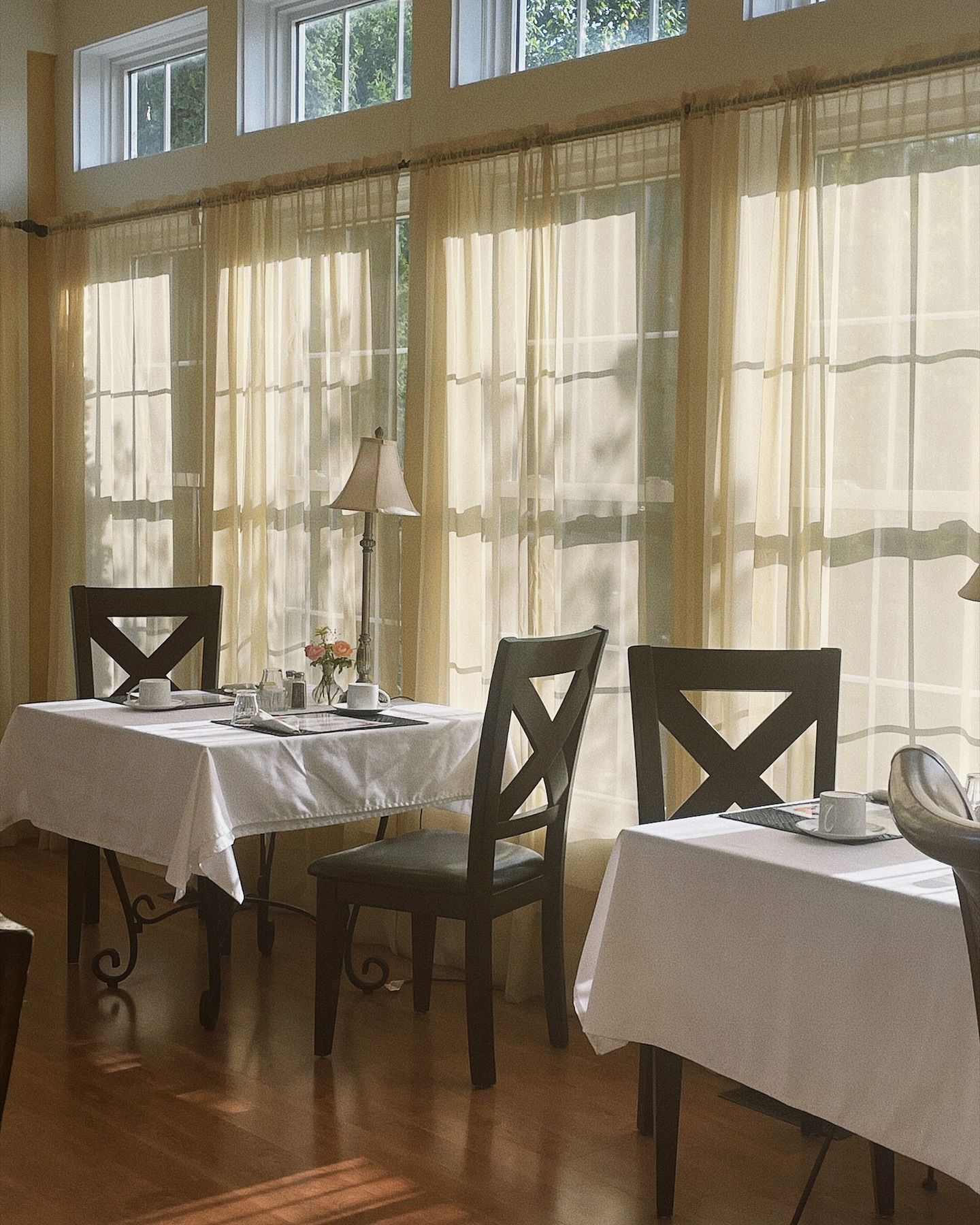 Sunlit room with two tables set for dining, sheer curtains, wood floors, and chairs.