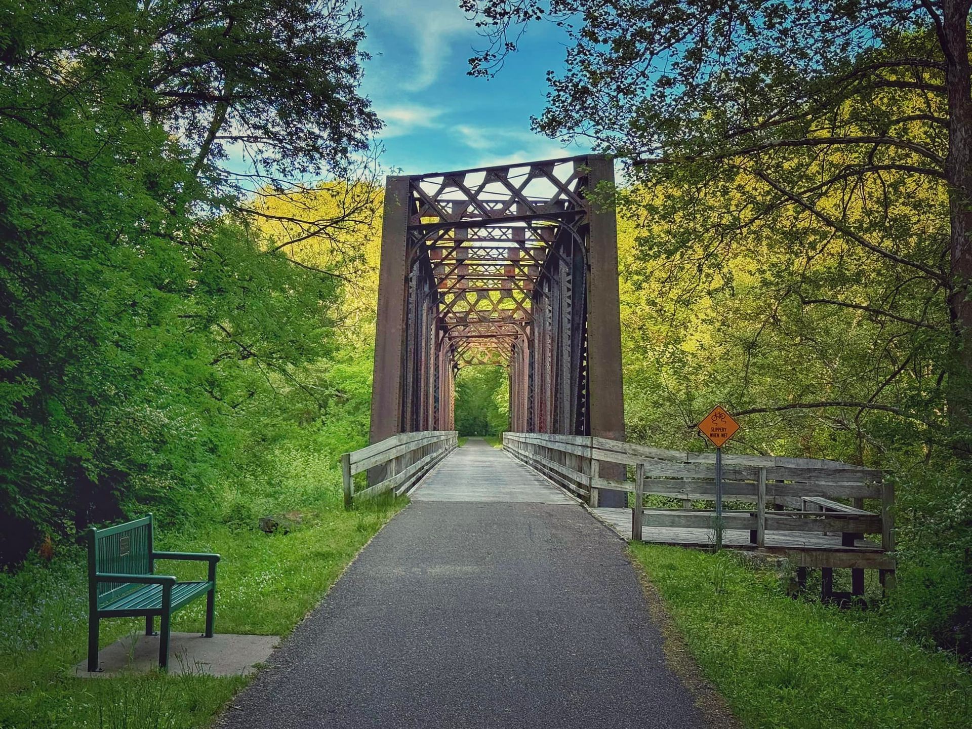 A paved trail leads to a historic steel truss bridge surrounded by lush green trees, with a green bench on the left.