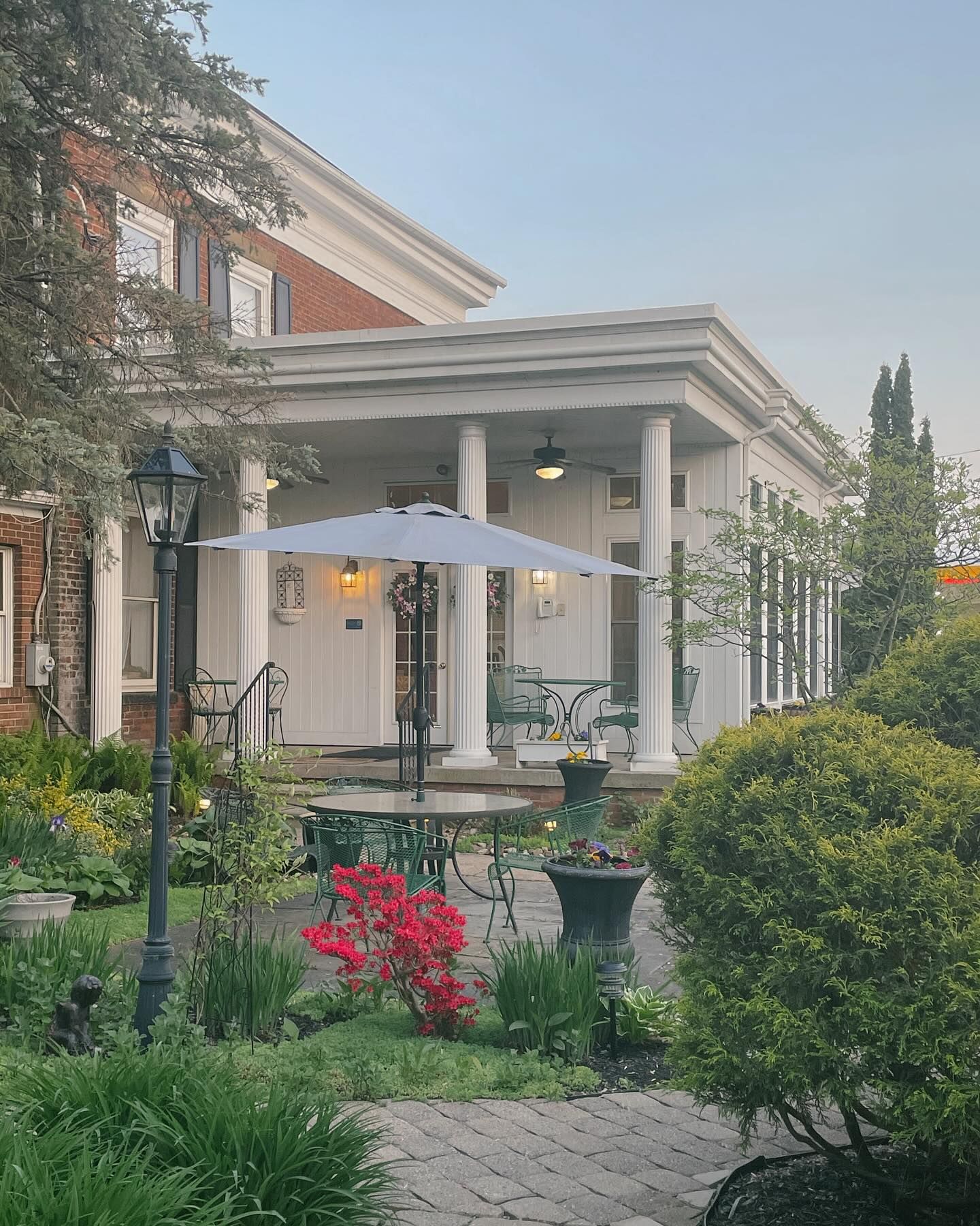 Exterior of a historic white building with columns. Outdoor seating area with umbrella and lush landscaping.
