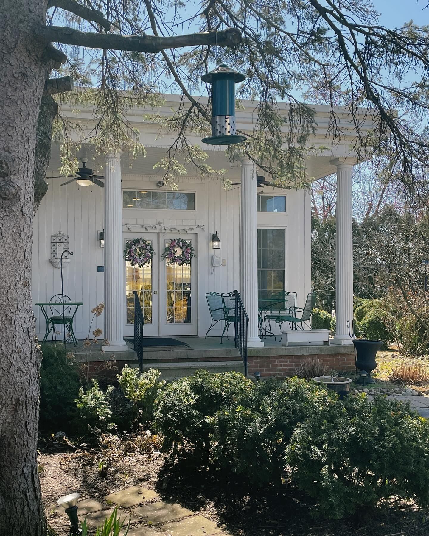White building with columns, double doors, and bird feeder. Green shrubs and outdoor seating visible.