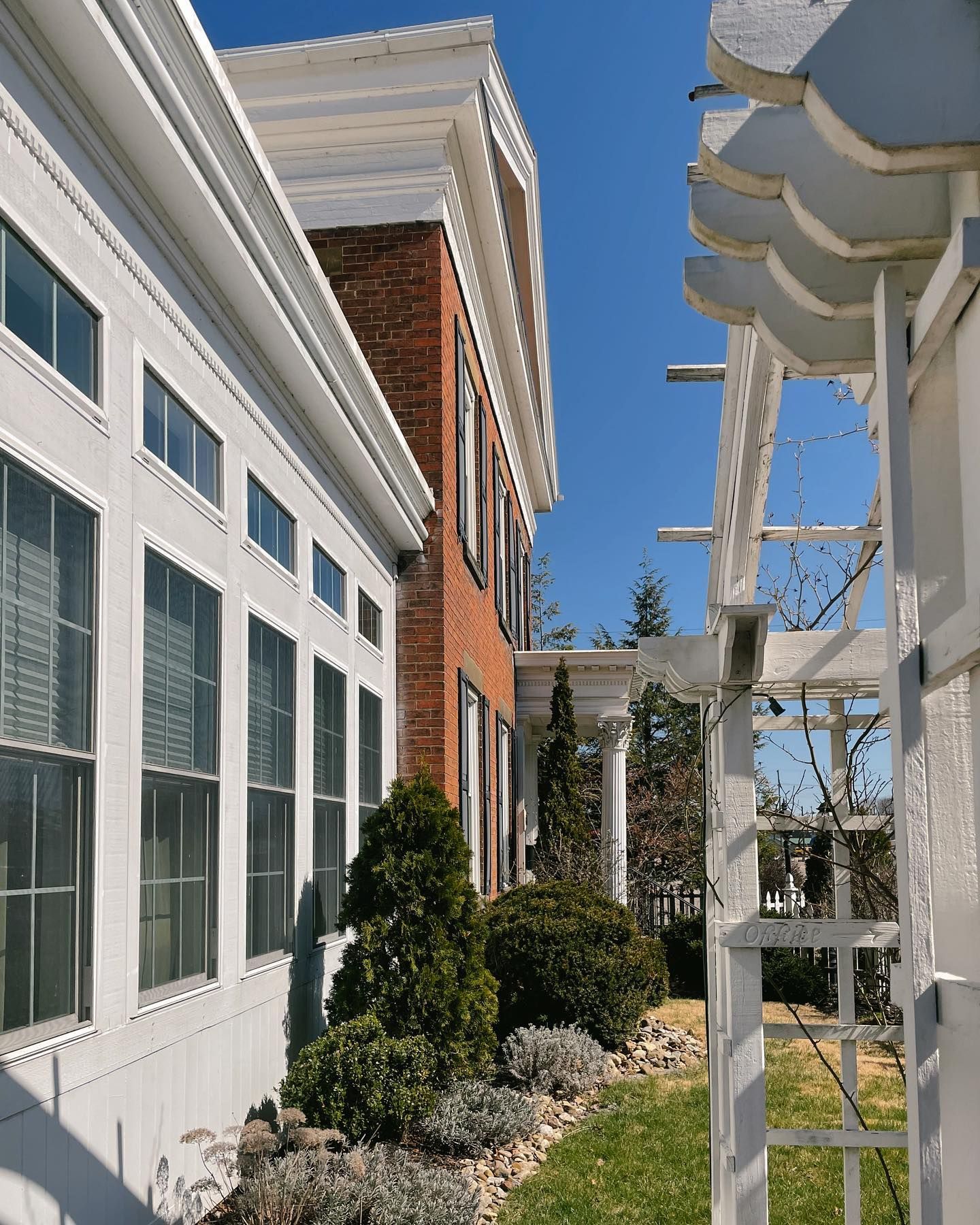 White house facade with multiple windows, next to a red brick building, and a white trellis under a blue sky.