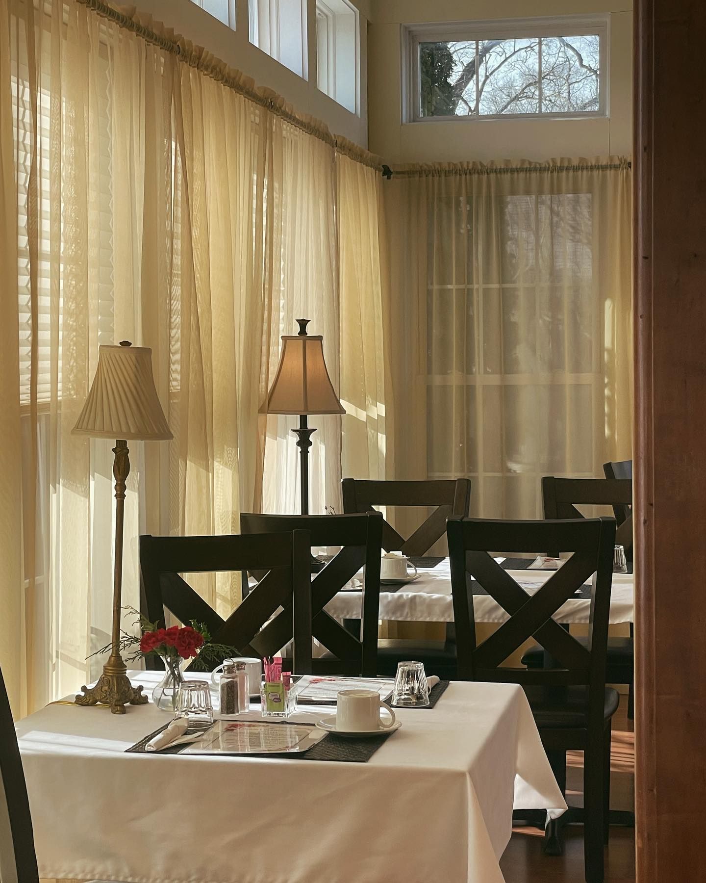 Dining room with white tablecloths, dark chairs, lamps, and sheer curtains filtering sunlight.