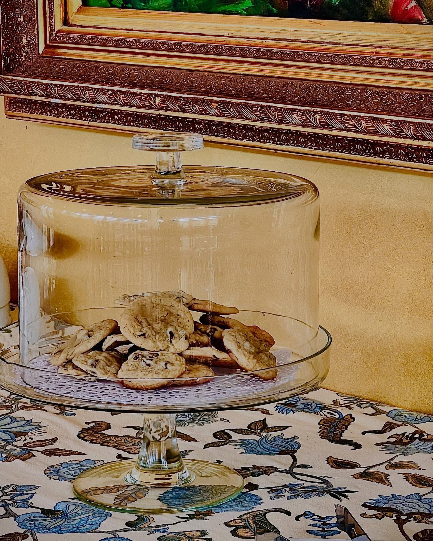 Cookies under a glass dome on a patterned tablecloth, with a painting in the background.