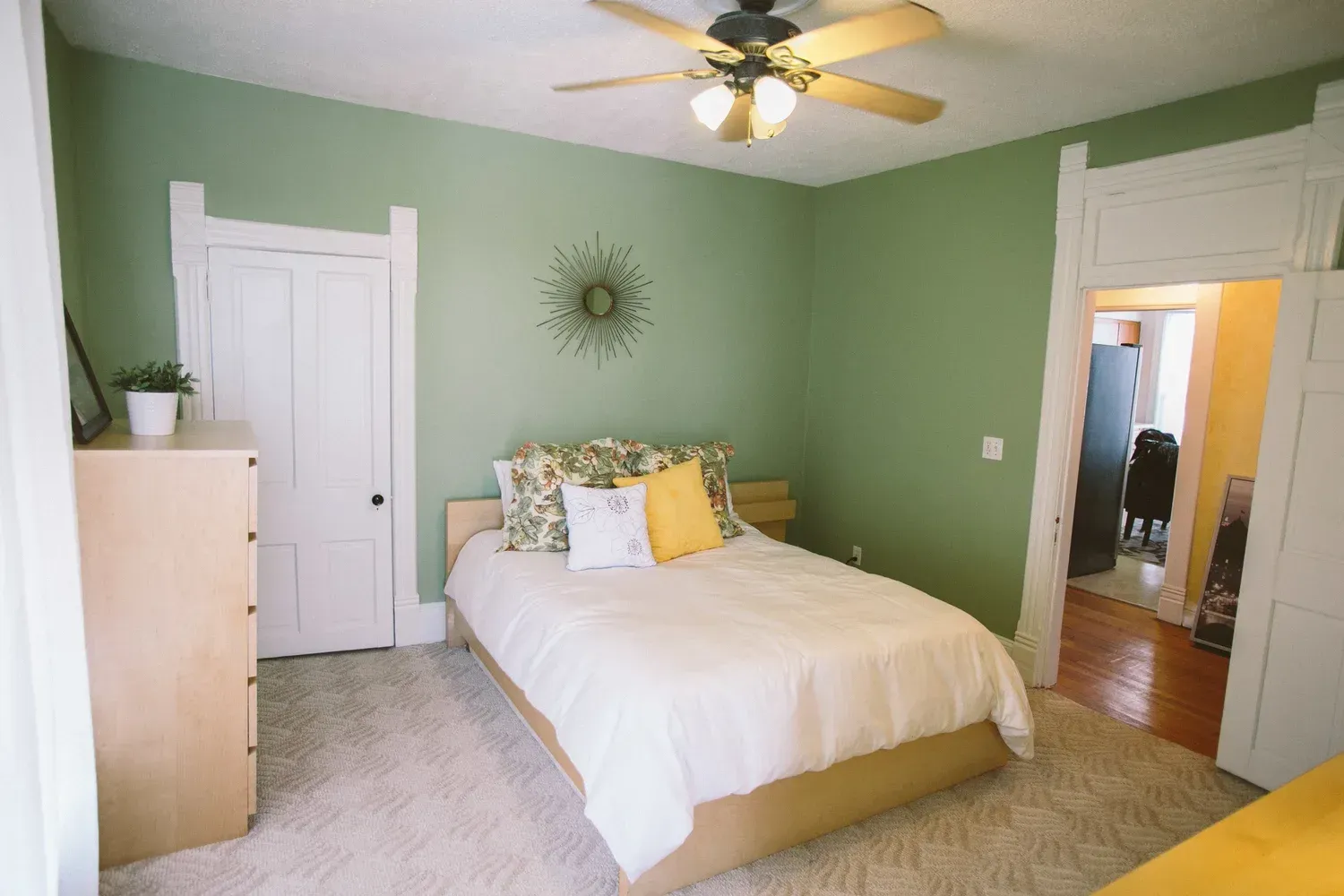 Green-walled bedroom with bed, white bedding, tan dresser, doorway, and ceiling fan.