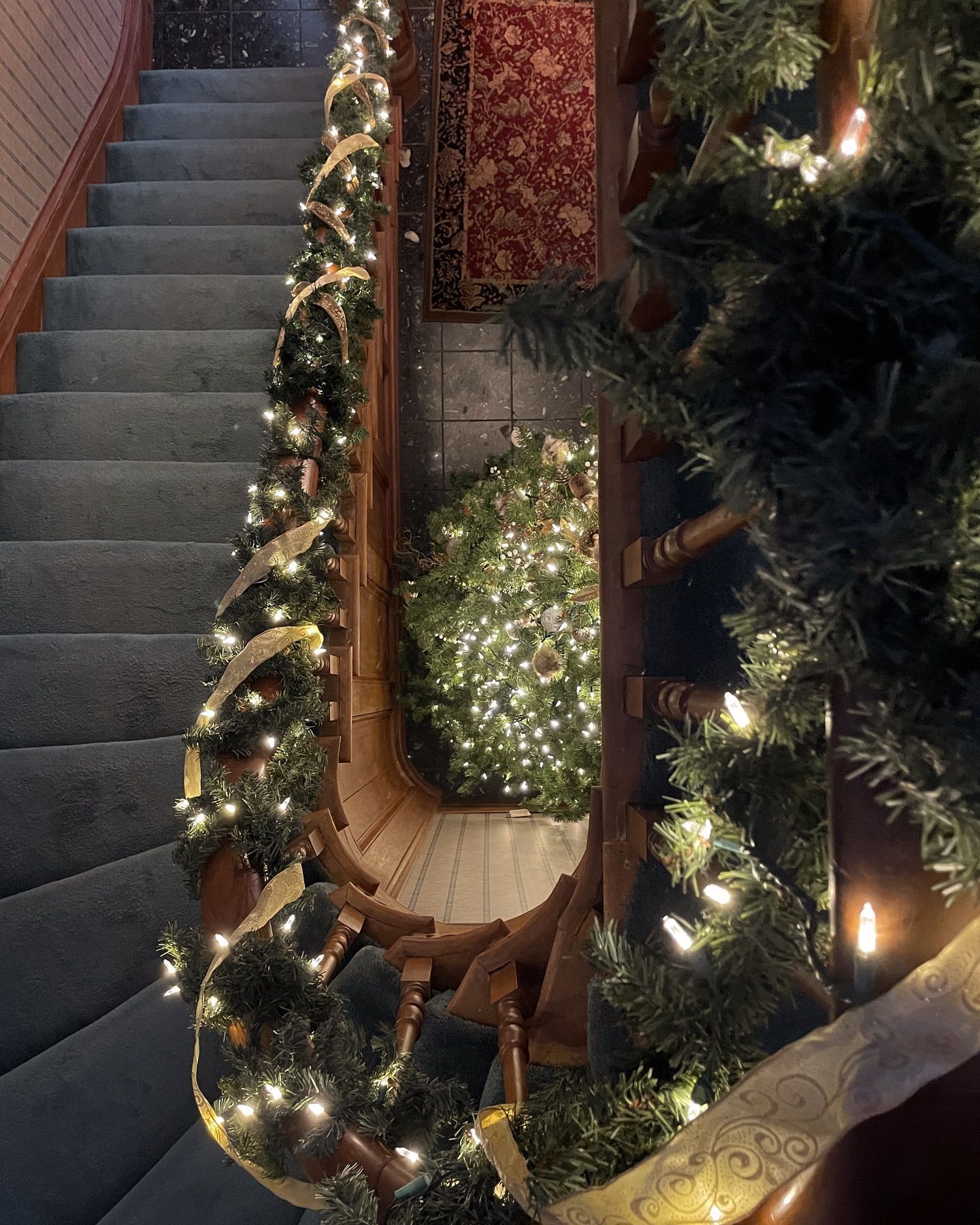 Staircase decorated with garlands, lights, and a small Christmas tree. Wooden railings and blue carpet.