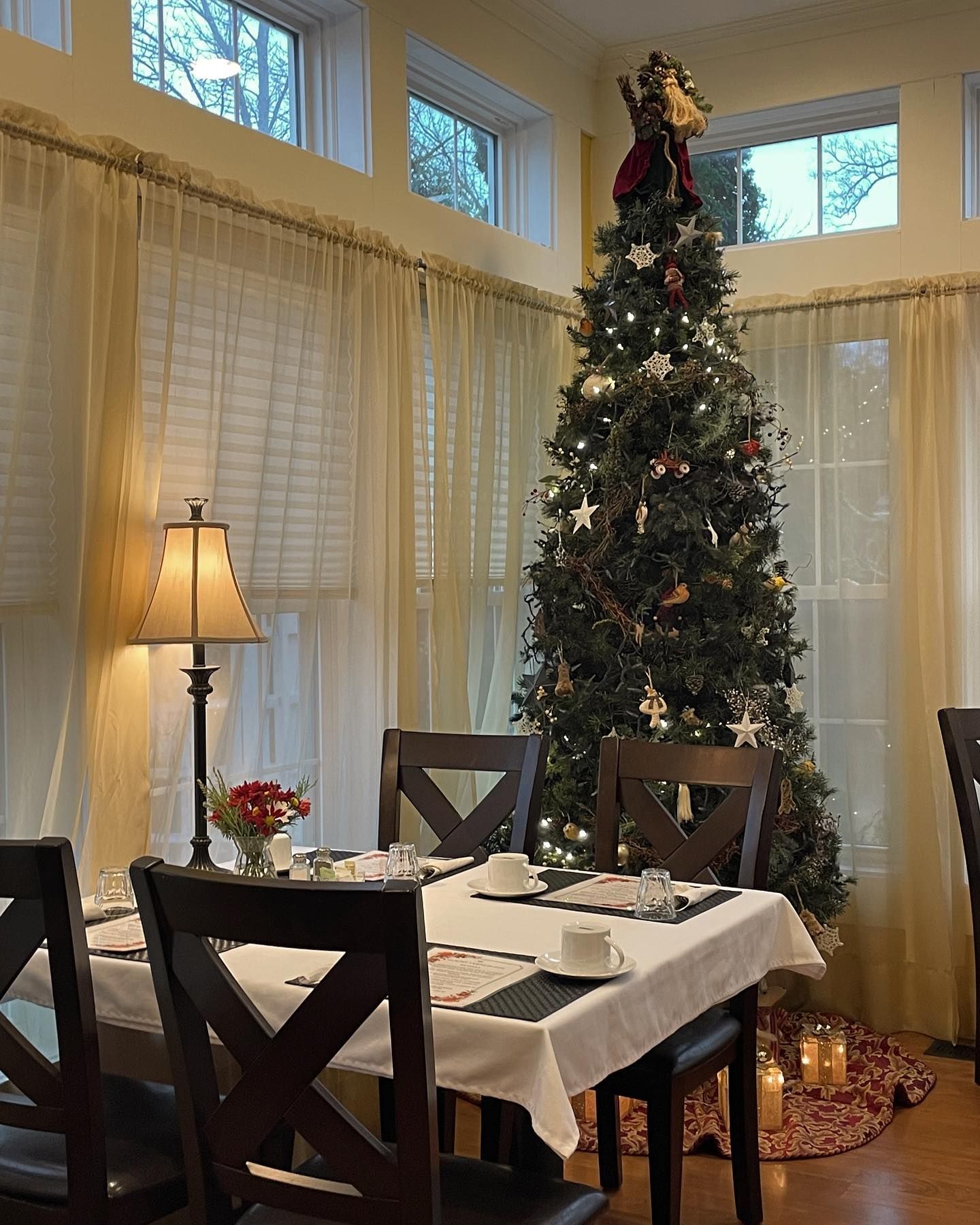 Dining room with decorated Christmas tree, table set with white tablecloth, and sheer curtains.