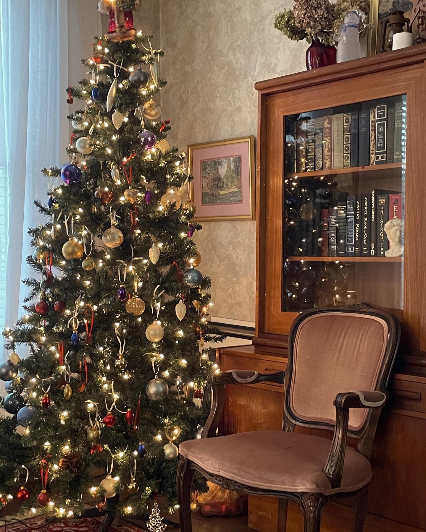 Christmas tree with ornaments, beside a cabinet with books and a velvet chair.