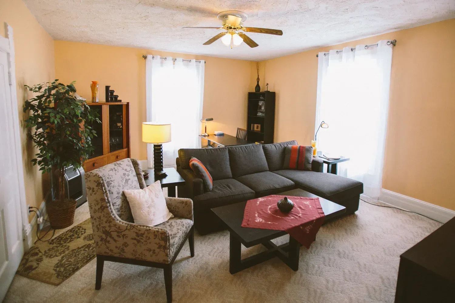 Living room with beige walls, a dark gray sectional sofa, and two windows with white curtains.
