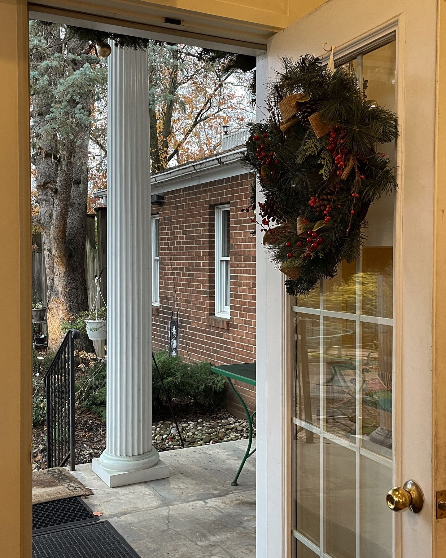 Open doorway with a wreath, leading to a porch with a column, brick building in the distance.