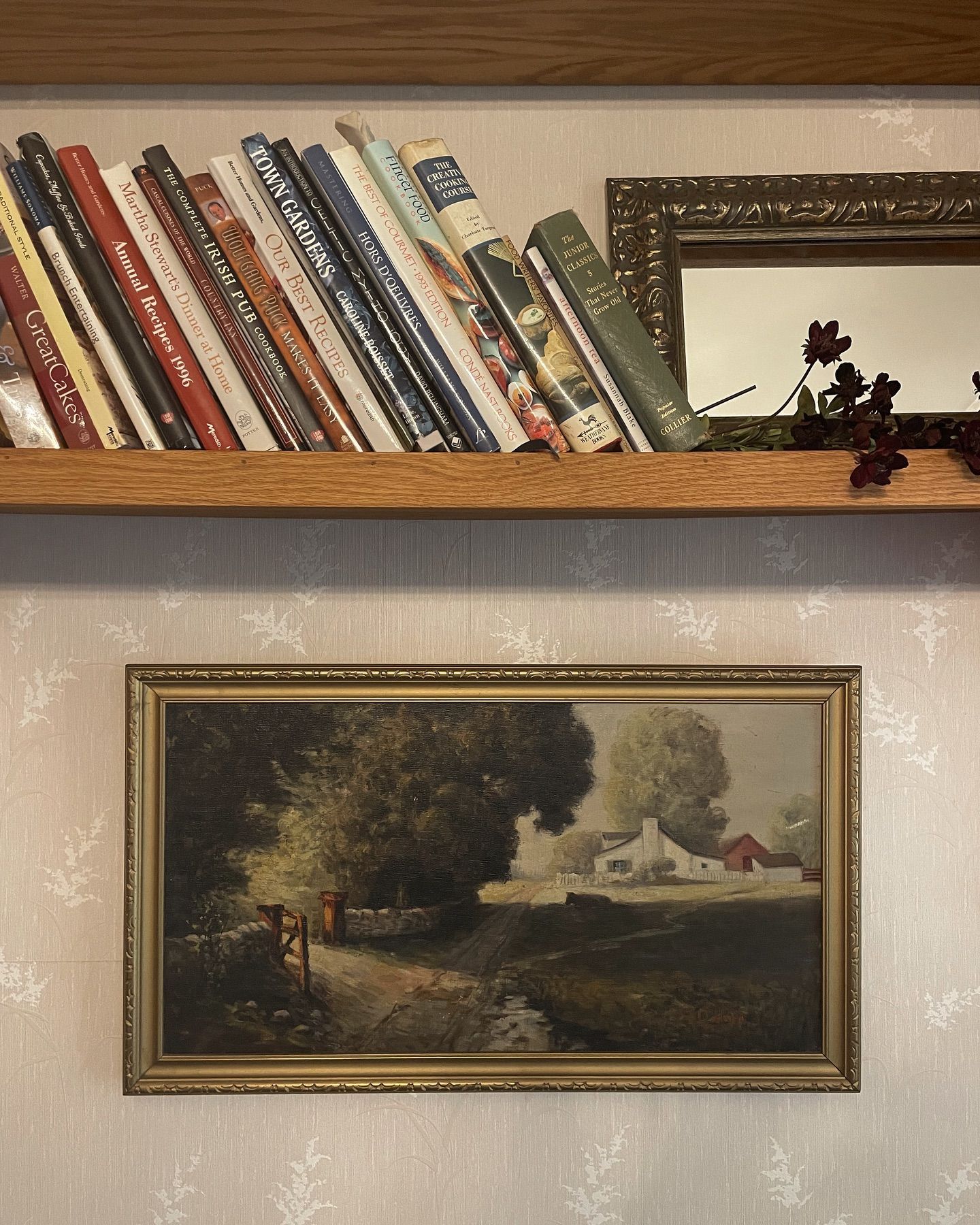 Books on shelf above framed landscape painting and small mirror. Wall is textured.
