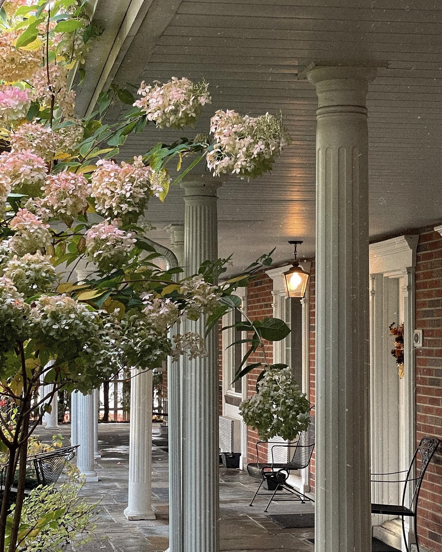 Stone columns support a porch with brick walls. Hydrangeas with pink and white blooms frame the view.