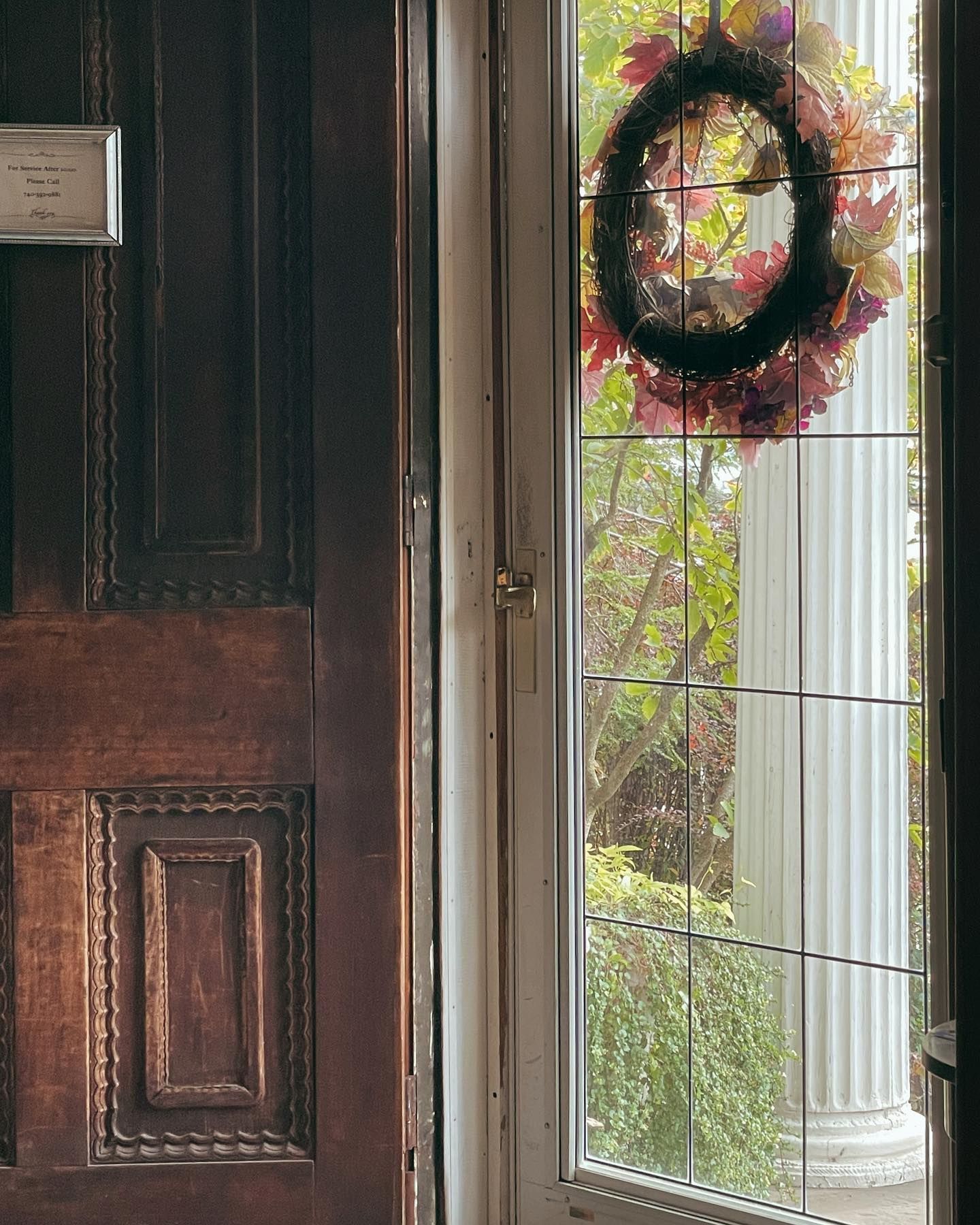 Dark wooden door next to a window with a wreath and a column visible, in an outdoor setting.