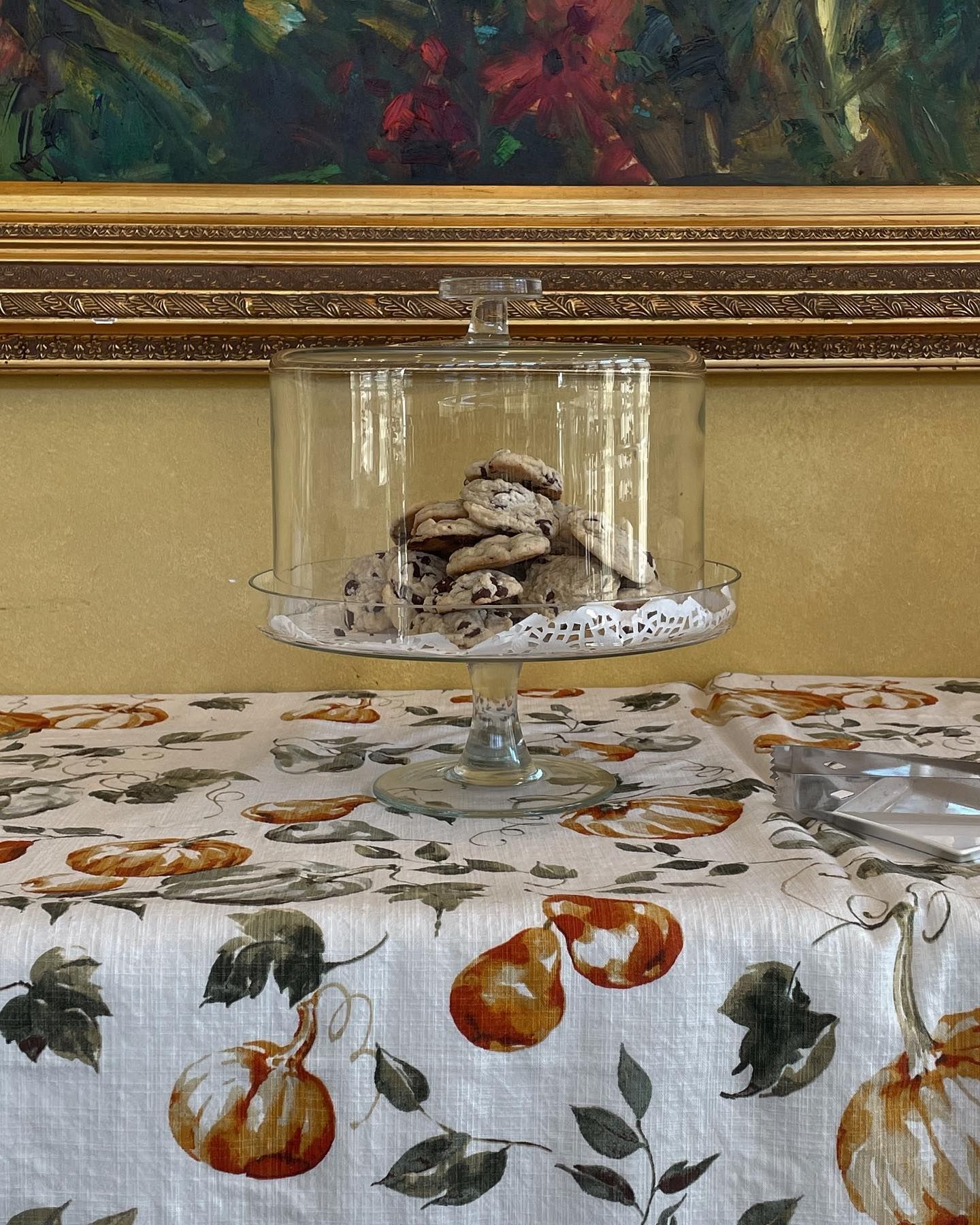 Cookies in a glass cake stand on a table with a pumpkin tablecloth, in front of a painting.